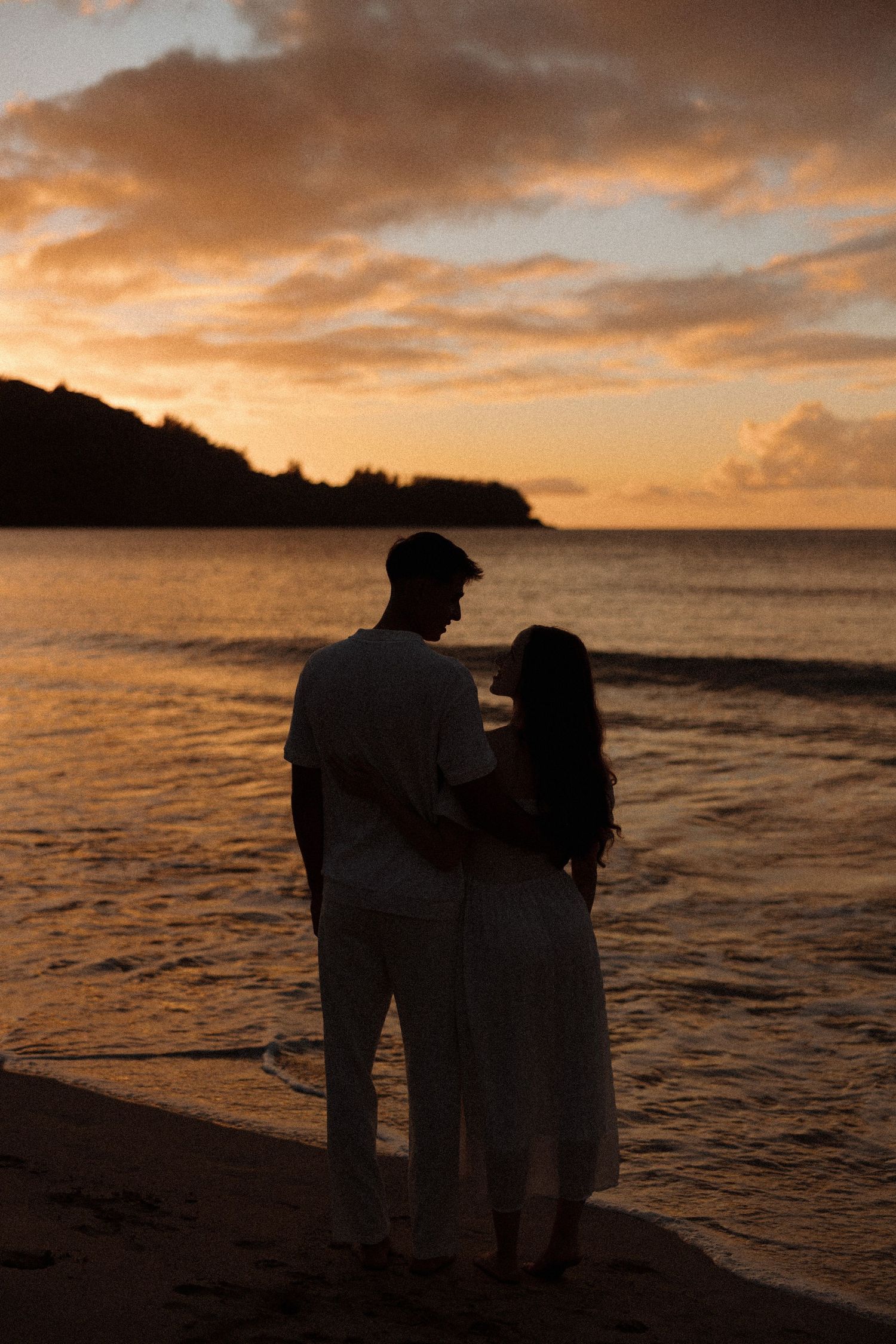 A romantic couple embraces on a beach at sunset with waves in the background.