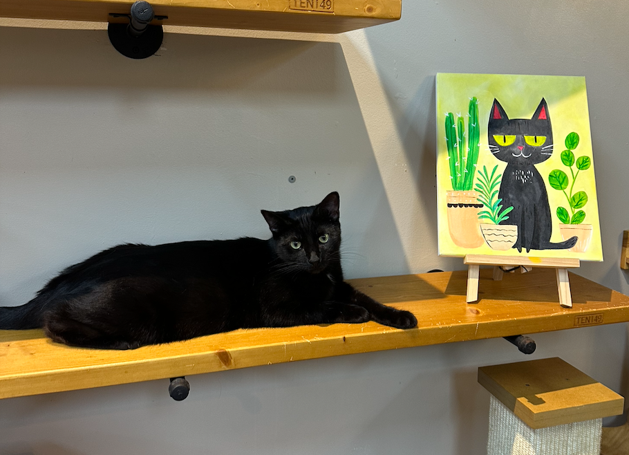 Black cat lounging on shelf next to its painted portrait featuring green plants.