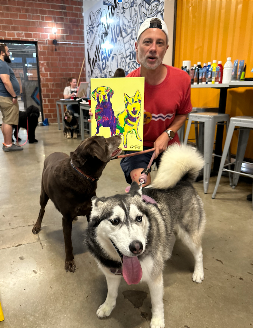 Two dogs posing with their owner who holds up a colorful pet portrait painting.