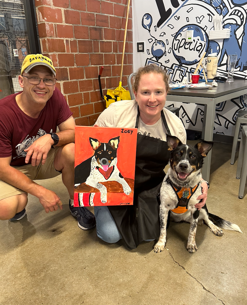 Two pet owners showing off their painted portrait next to their spotted dog.