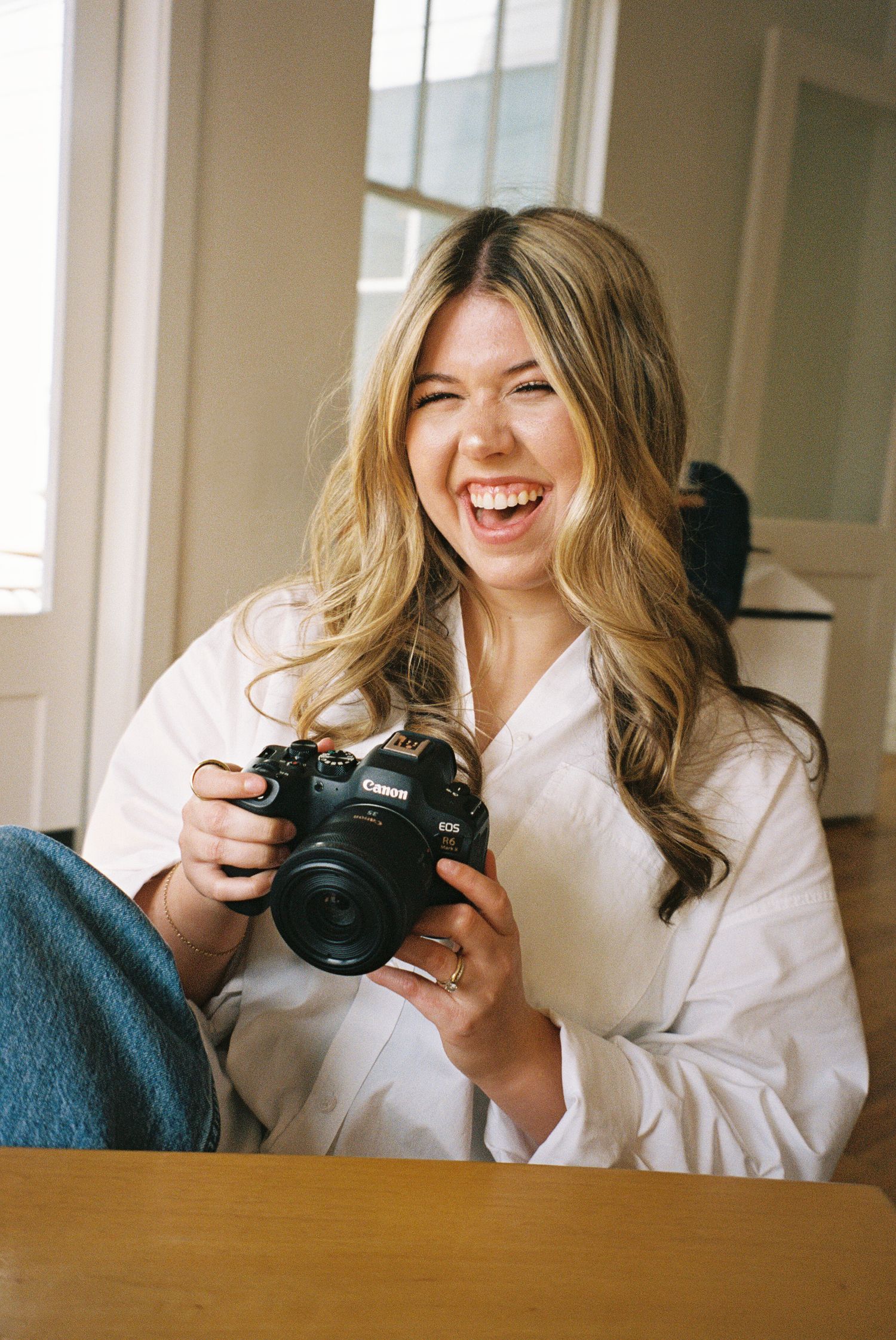 Laughing blonde woman in white shirt holding a Canon EOS camera while sitting indoors near a window.