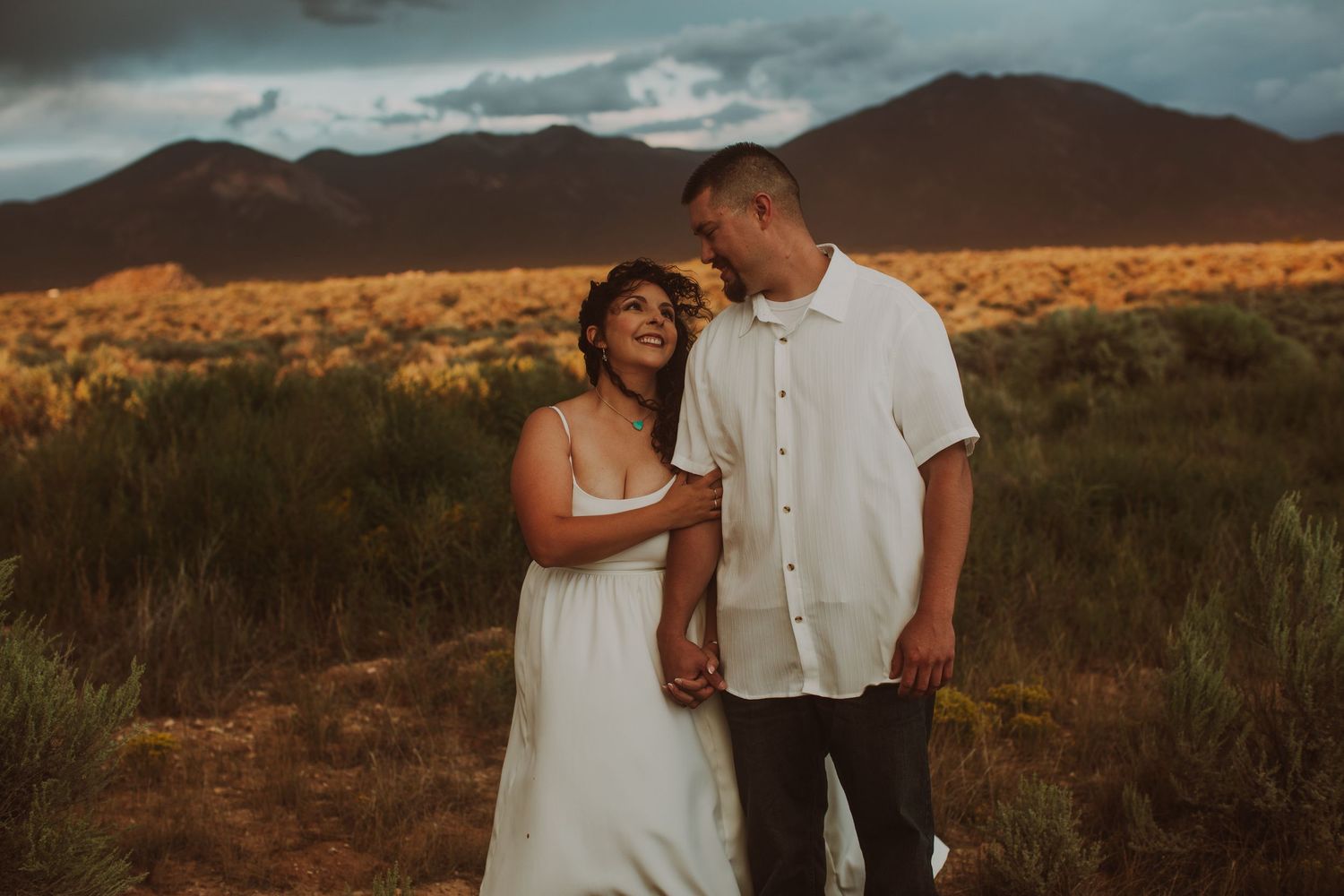 Couple in white outfits laughing together in golden desert field with dramatic mountain backdrop at sunset.