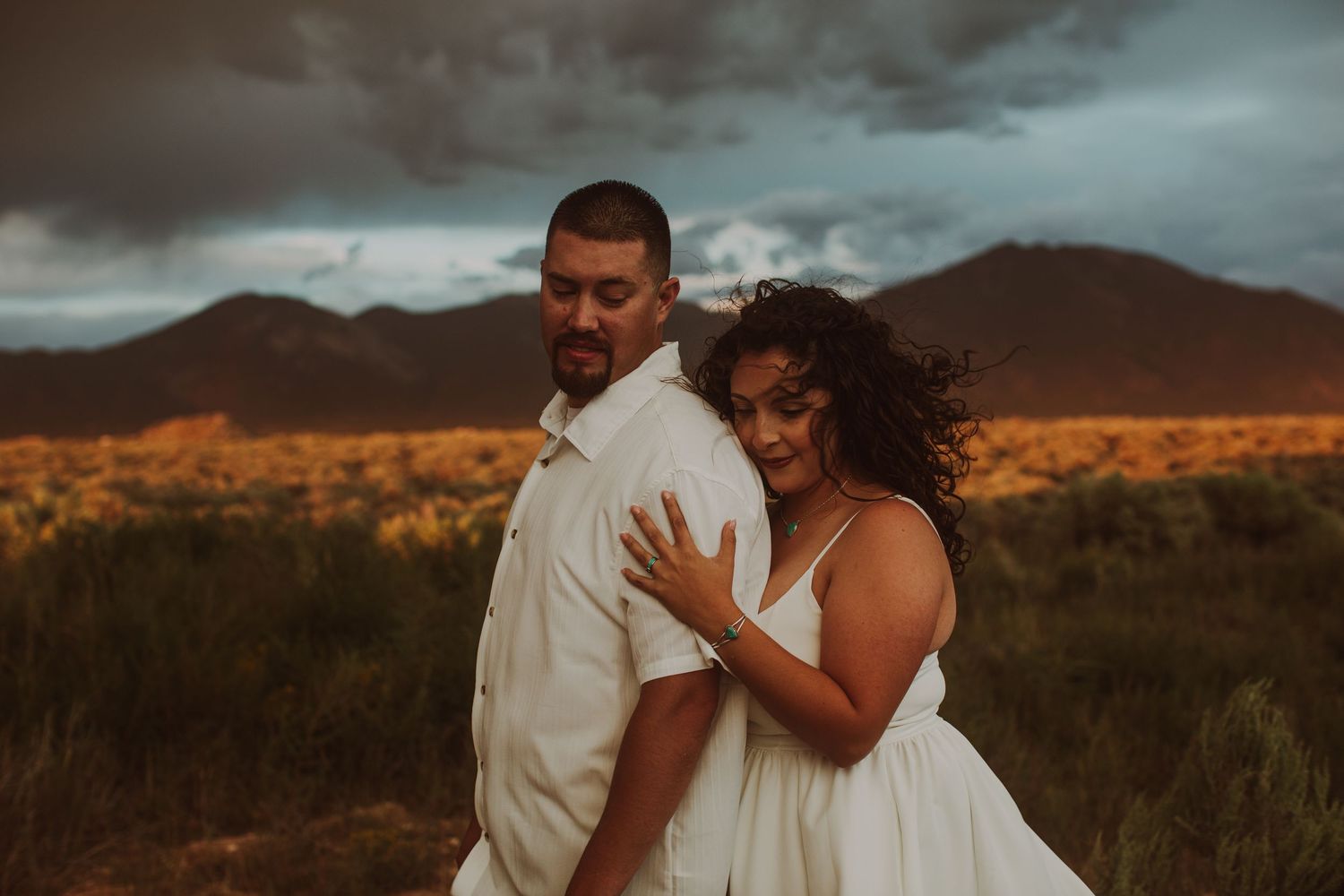 Romantic couple embracing in desert landscape with stormy sky and red mountains during golden hour shoot.