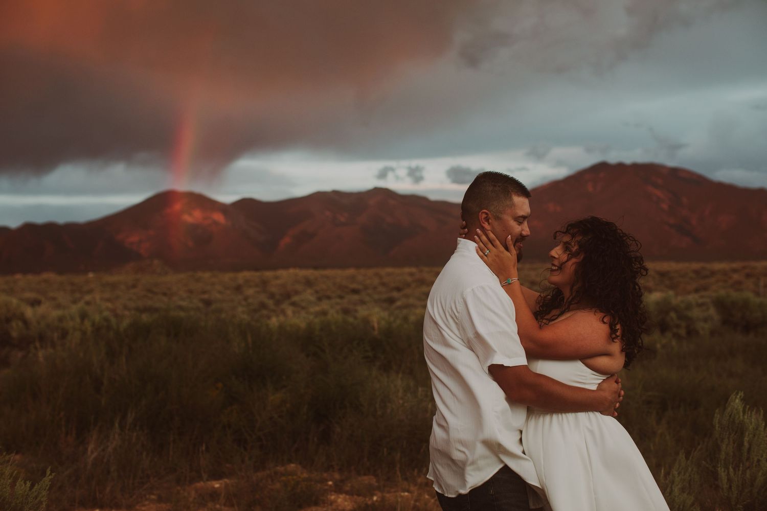 Couple sharing intimate moment in desert field with vibrant rainbow and red-hued mountains at sunset.