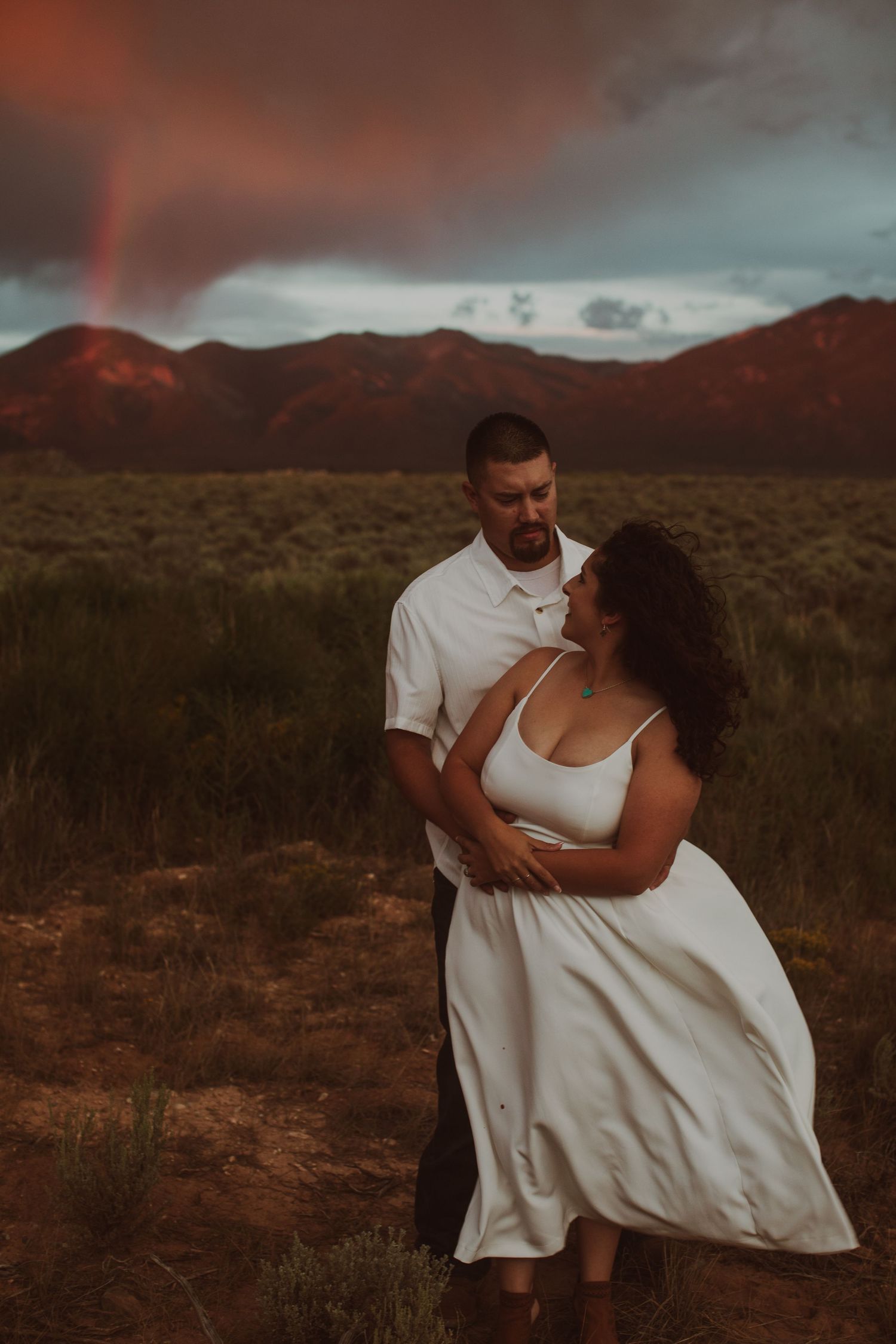 Couple in white attire sharing tender embrace in desert field under dramatic red stormy sunset sky.