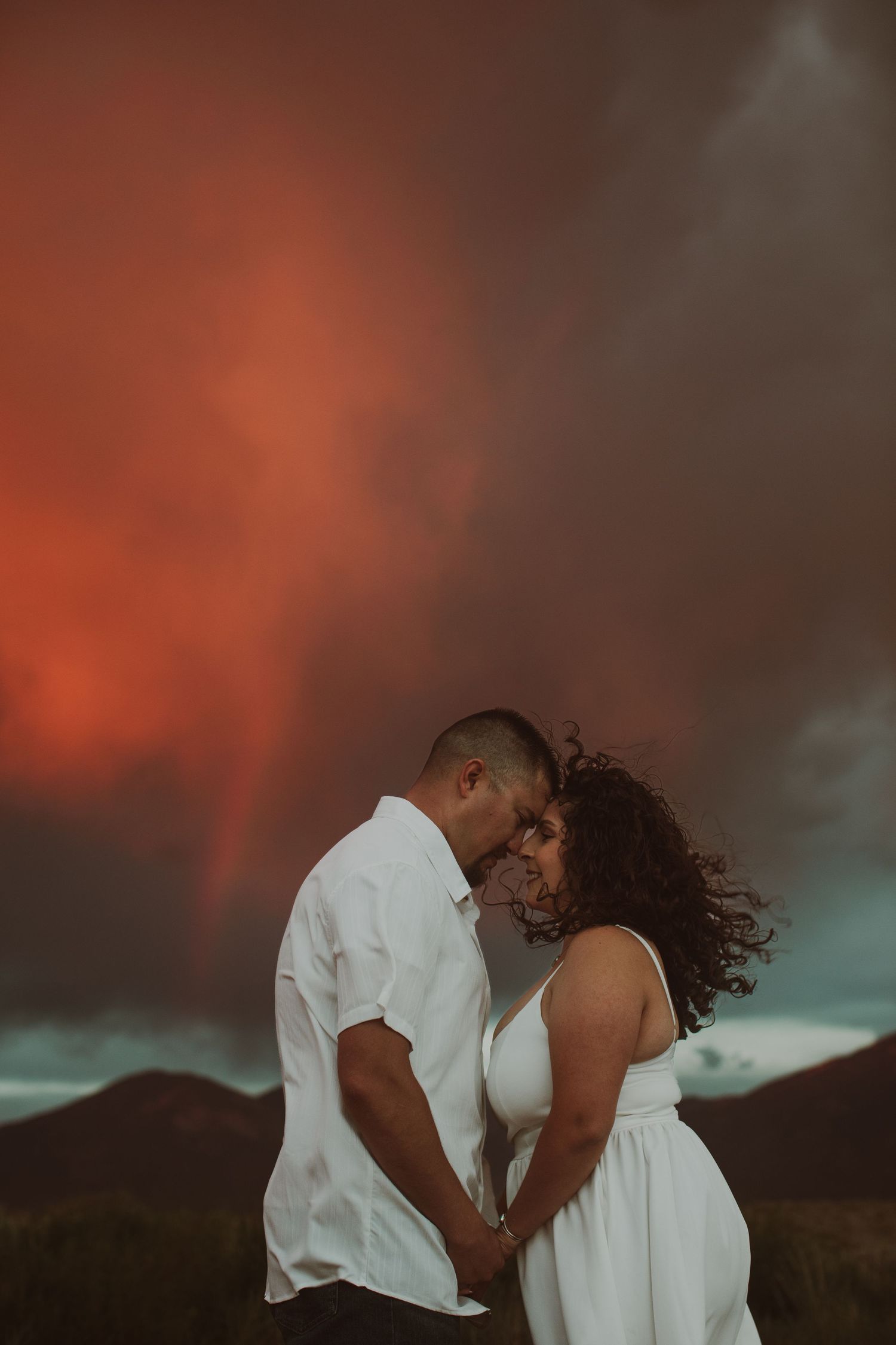 Romantic couple holding hands forehead-to-forehead under vivid red stormy sky with rainbow in background.