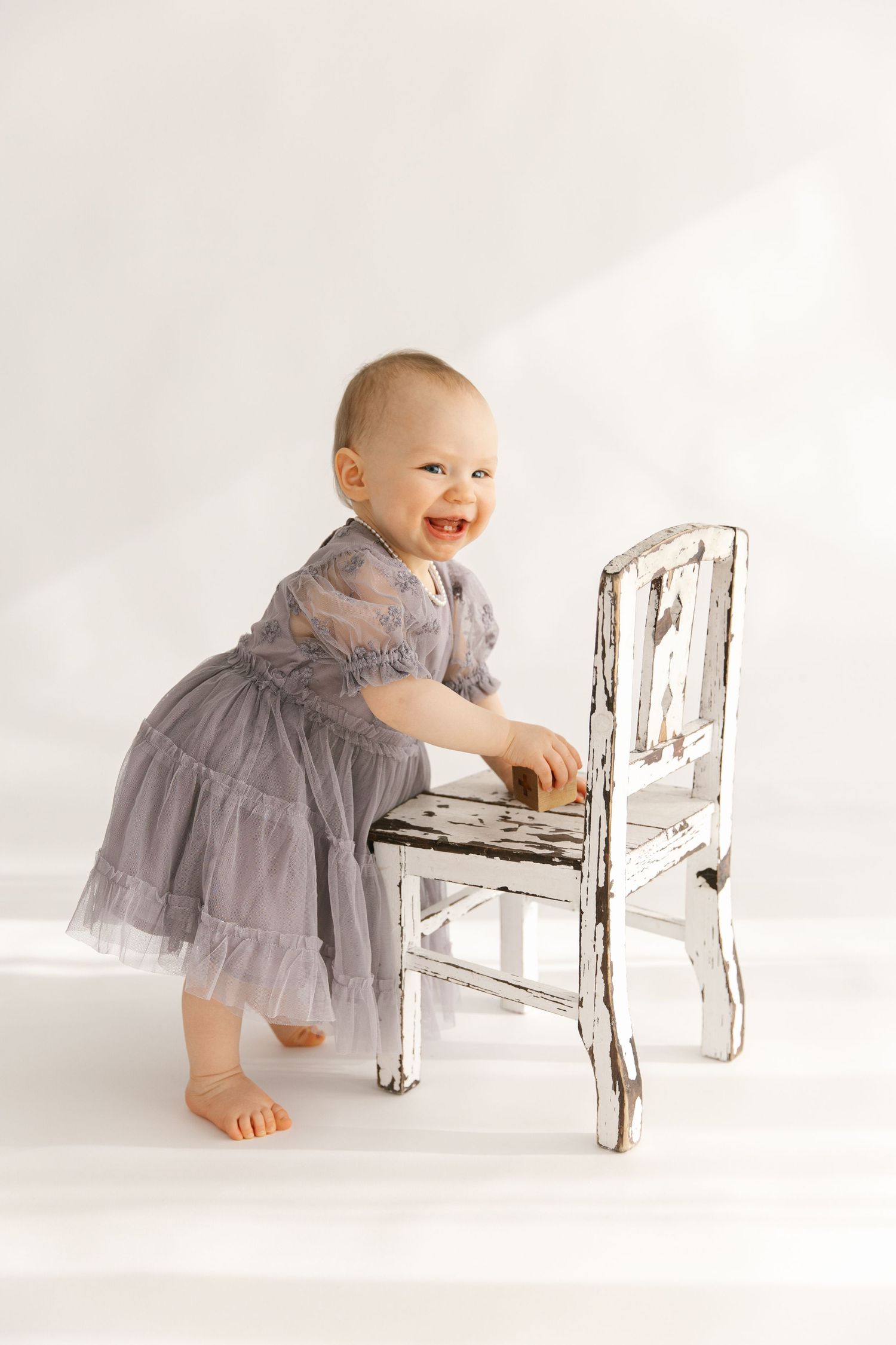 Baby in a gray dress smiles while leaning on a distressed white wooden chair in a minimalist studio setting.