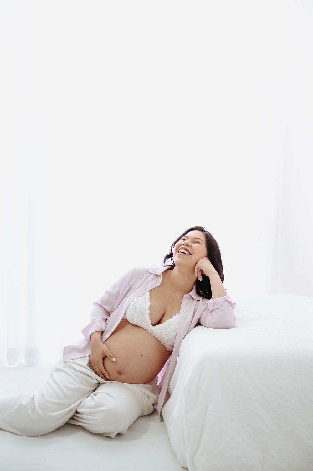 Smiling pregnant woman in pink shirt and lace bra sitting on floor, laughing while talking on phone.
