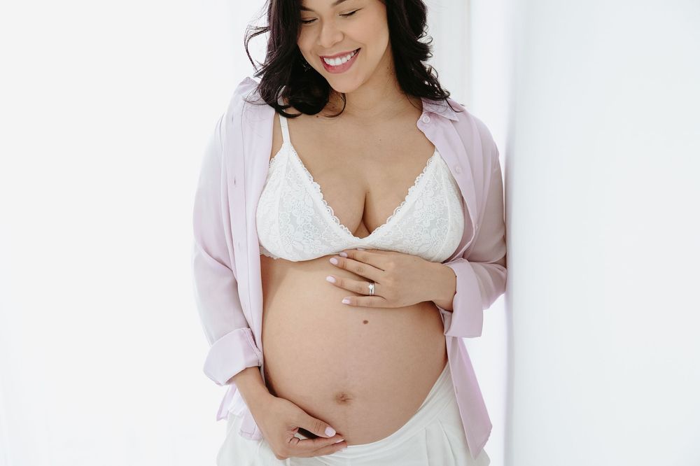 Smiling pregnant woman in white lace bra and pink robe cradling her baby bump against a white background.