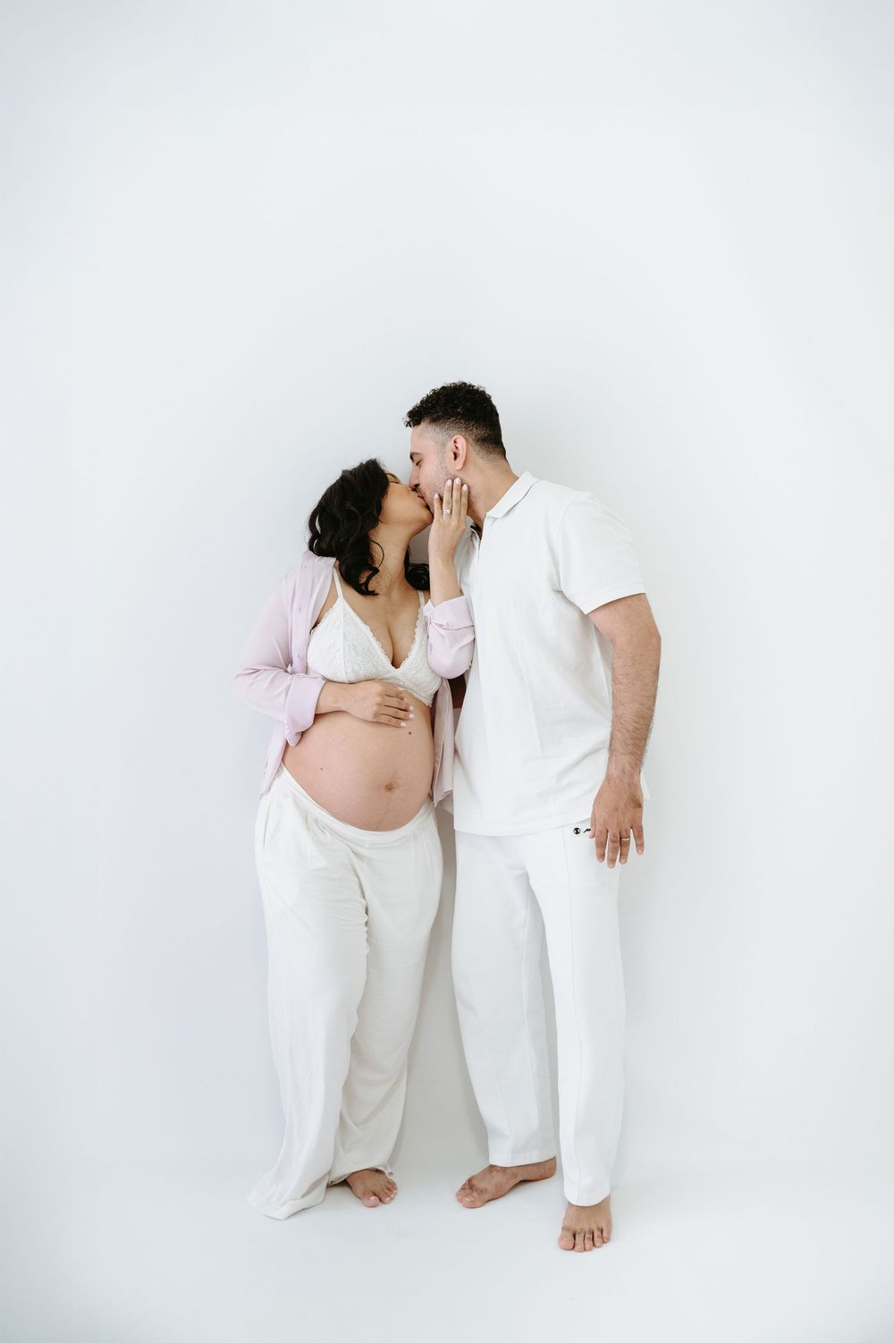 Pregnant couple sharing a tender kiss, woman in pink top and white pants, man in white outfit, against white background.