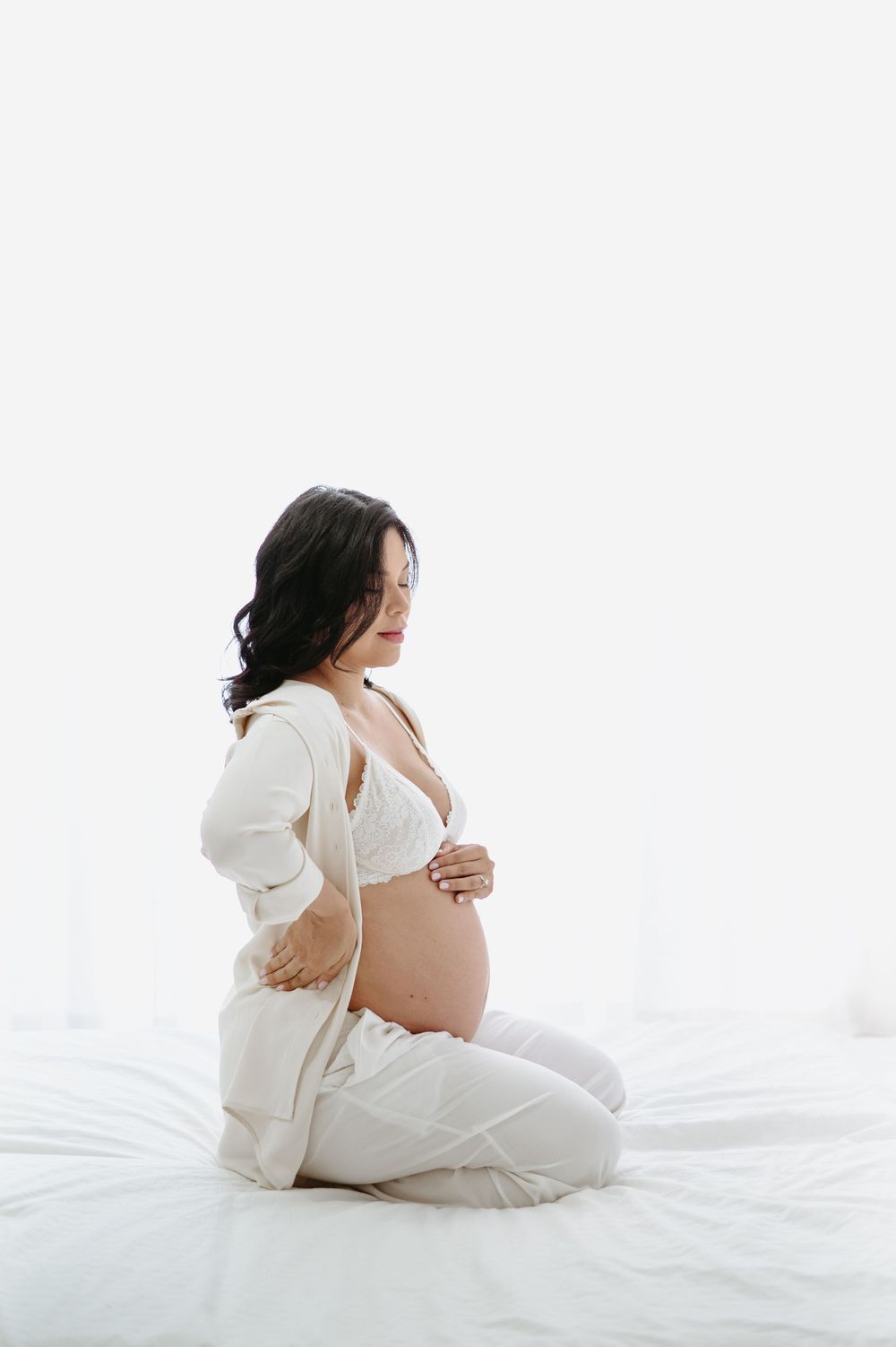 Pregnant woman in white lingerie and cardigan sitting on bed cradling her baby bump in a serene maternity photo.