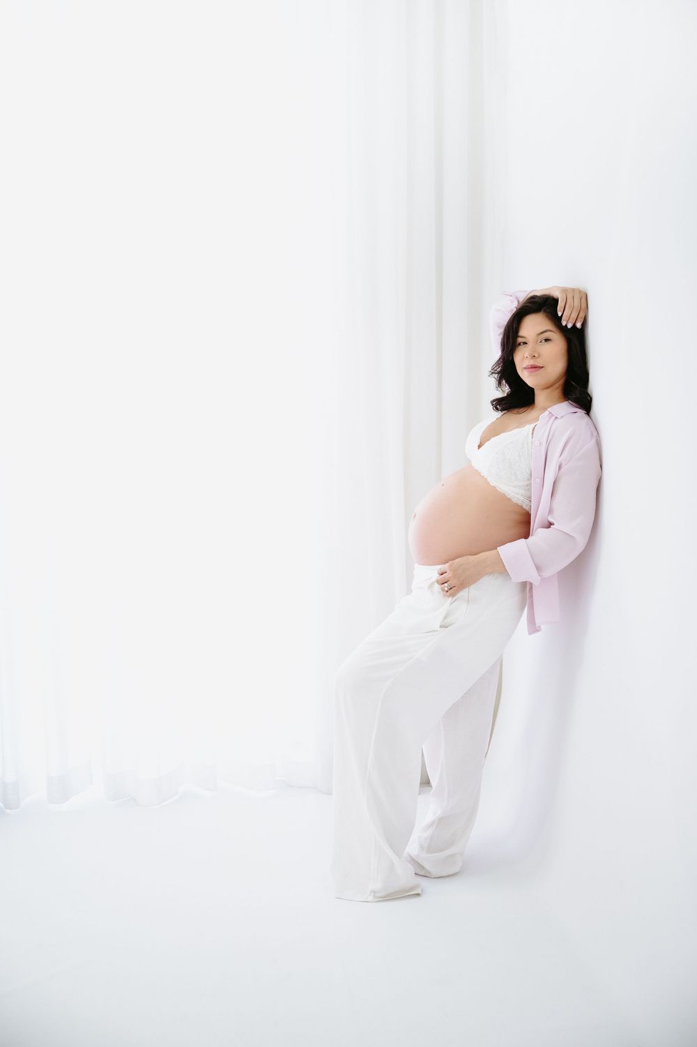 Pregnant woman in white outfit and pink cardigan leaning against white wall, cradling baby bump in bright studio.