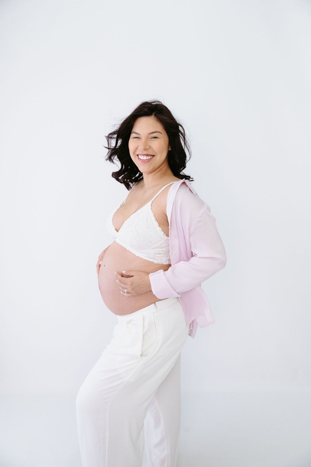 Smiling pregnant woman in white bra and pink cardigan holding her baby bump against a white background.