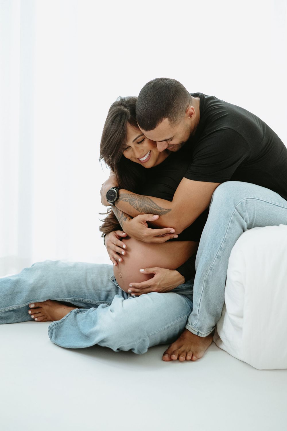 Happy couple sharing a tender moment, man embracing pregnant woman sitting on white floor, both smiling and touching baby bump.