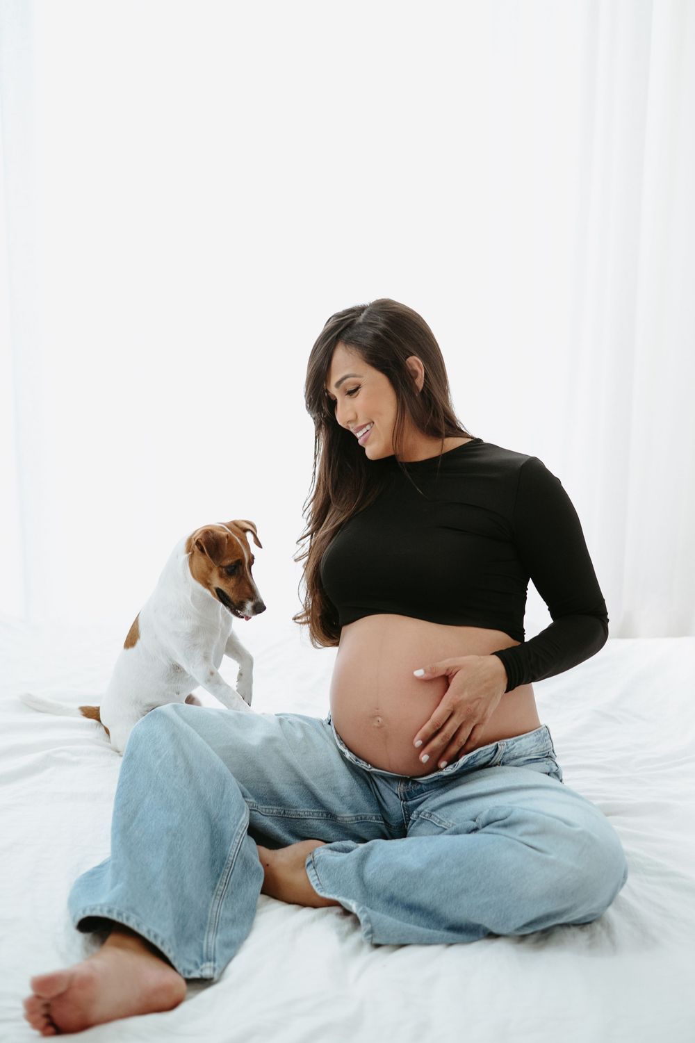 Pregnant woman in black crop top and jeans sits on bed smiling at her bump, with a small Jack Russell terrier dog beside her.