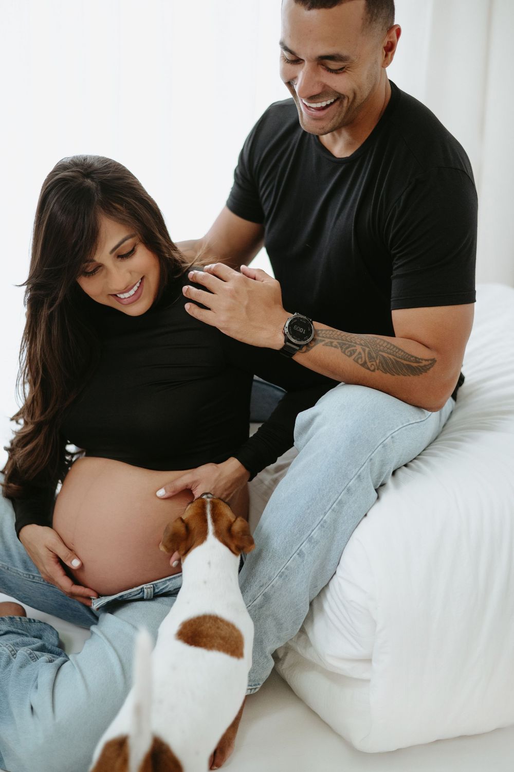 Smiling pregnant couple in black tops with a Jack Russell terrier dog sitting on a white bed.