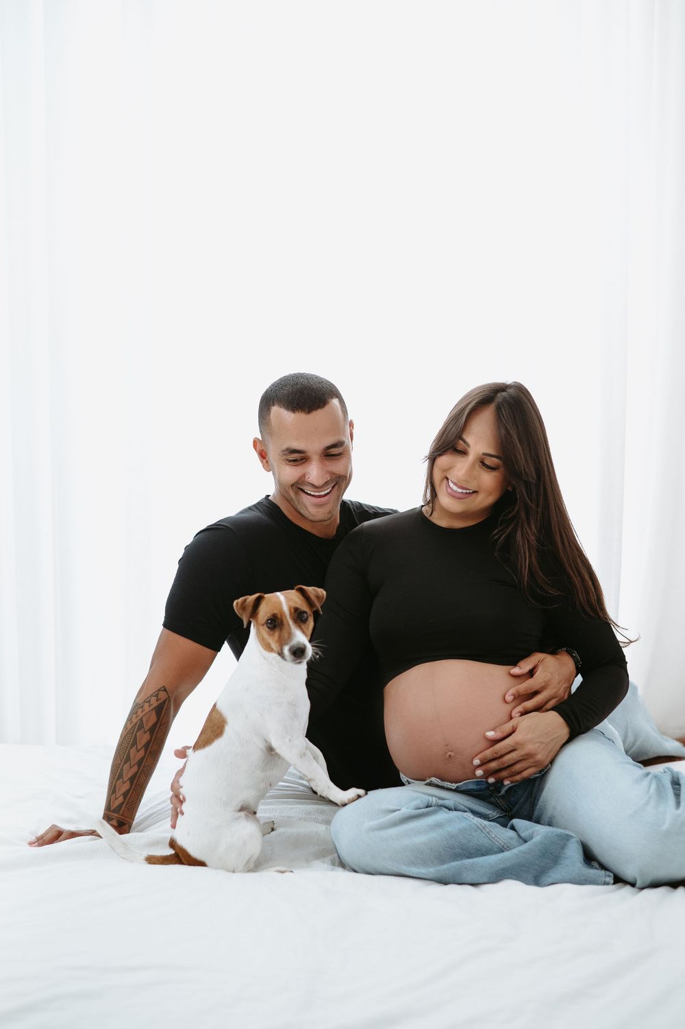 Smiling couple in black tops sitting on bed with Jack Russell terrier, pregnant woman cradling baby bump.