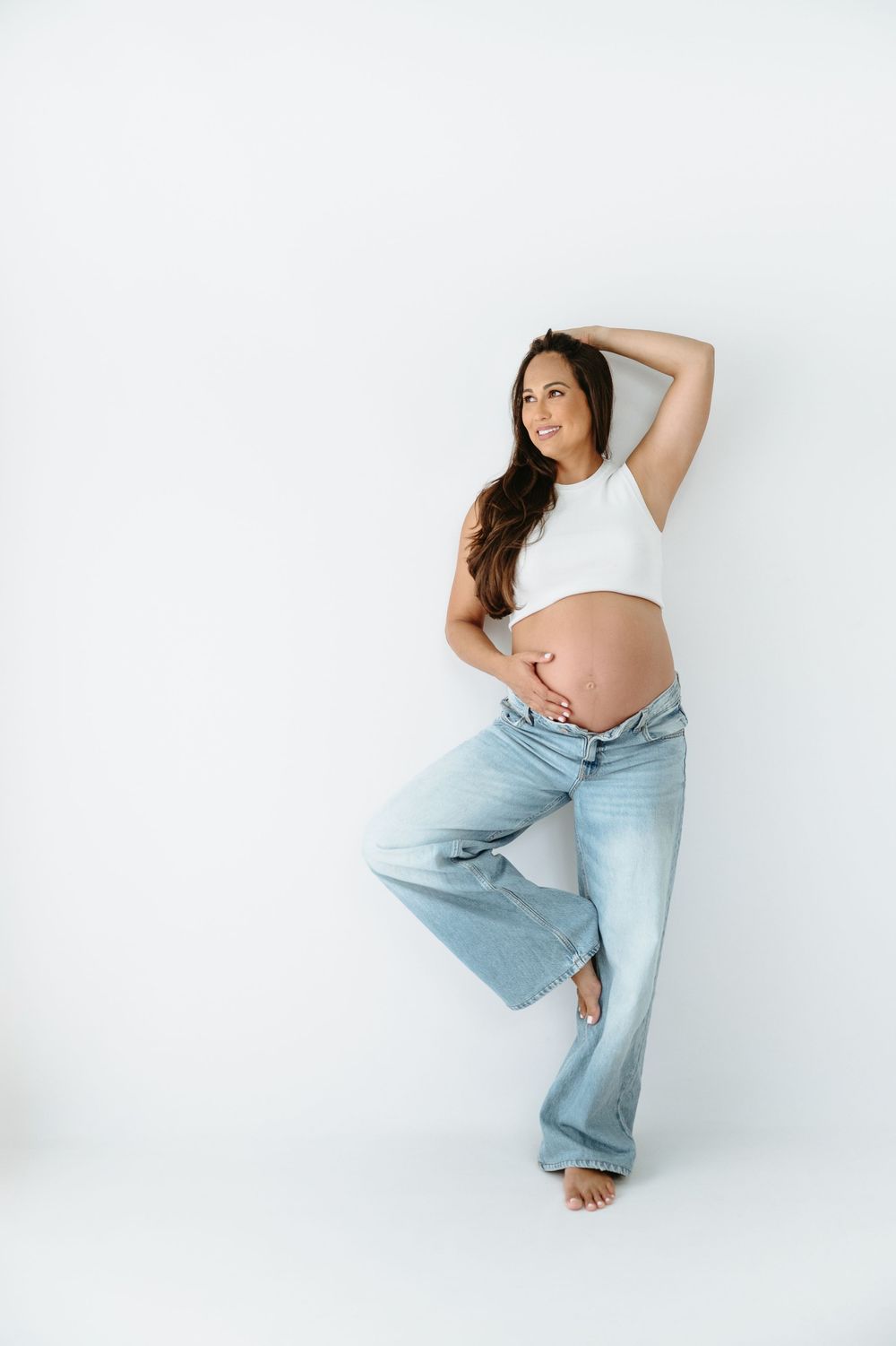 Pregnant woman in white crop top and light blue flare jeans posing against white background, holding baby bump.