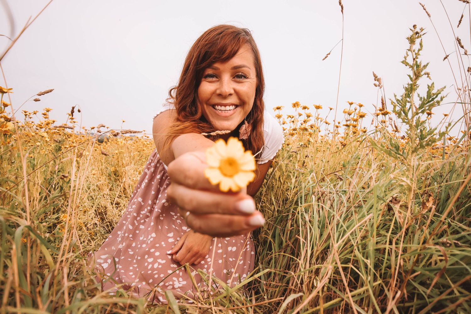 Person in white dress and brown hat sitting among tall grass during golden hour.