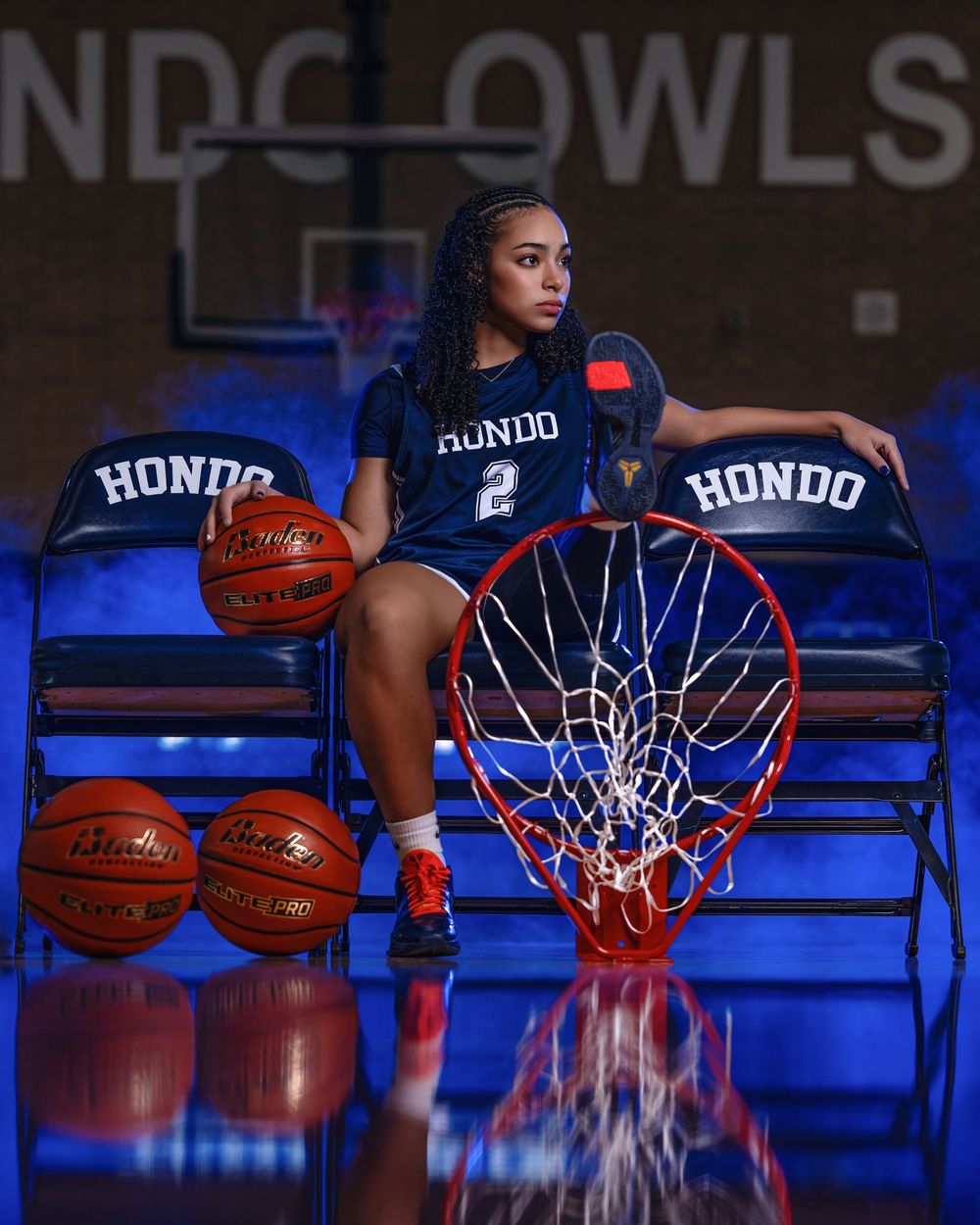 Basketball player in navy Hondo jersey number 2 poses on bleachers with basketballs and hoop in dramatic lighting setup.
