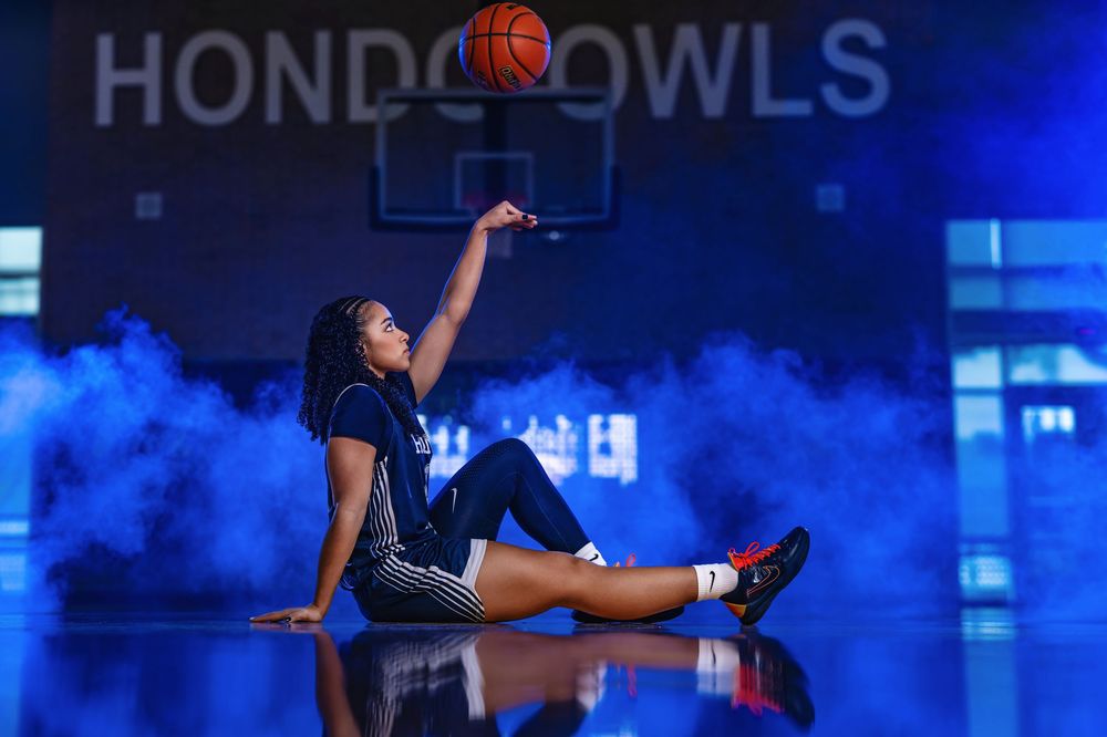 Basketball player in blue jersey dribbles against dramatic blue lighting during indoor game.