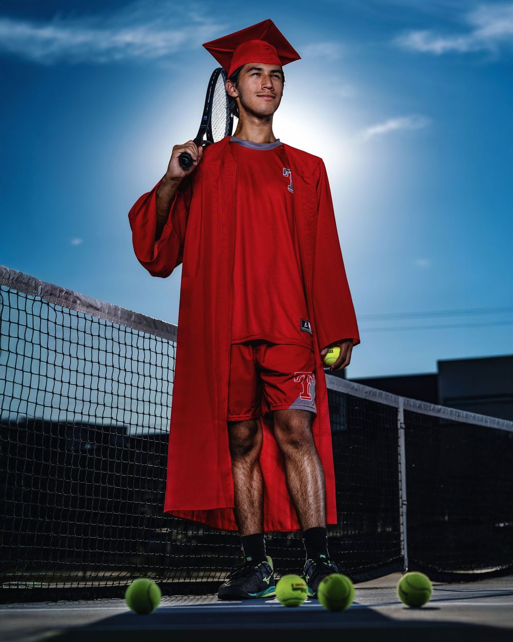 Graduate in red cap and gown stands on tennis court with tennis balls scattered around during sunset photo session.