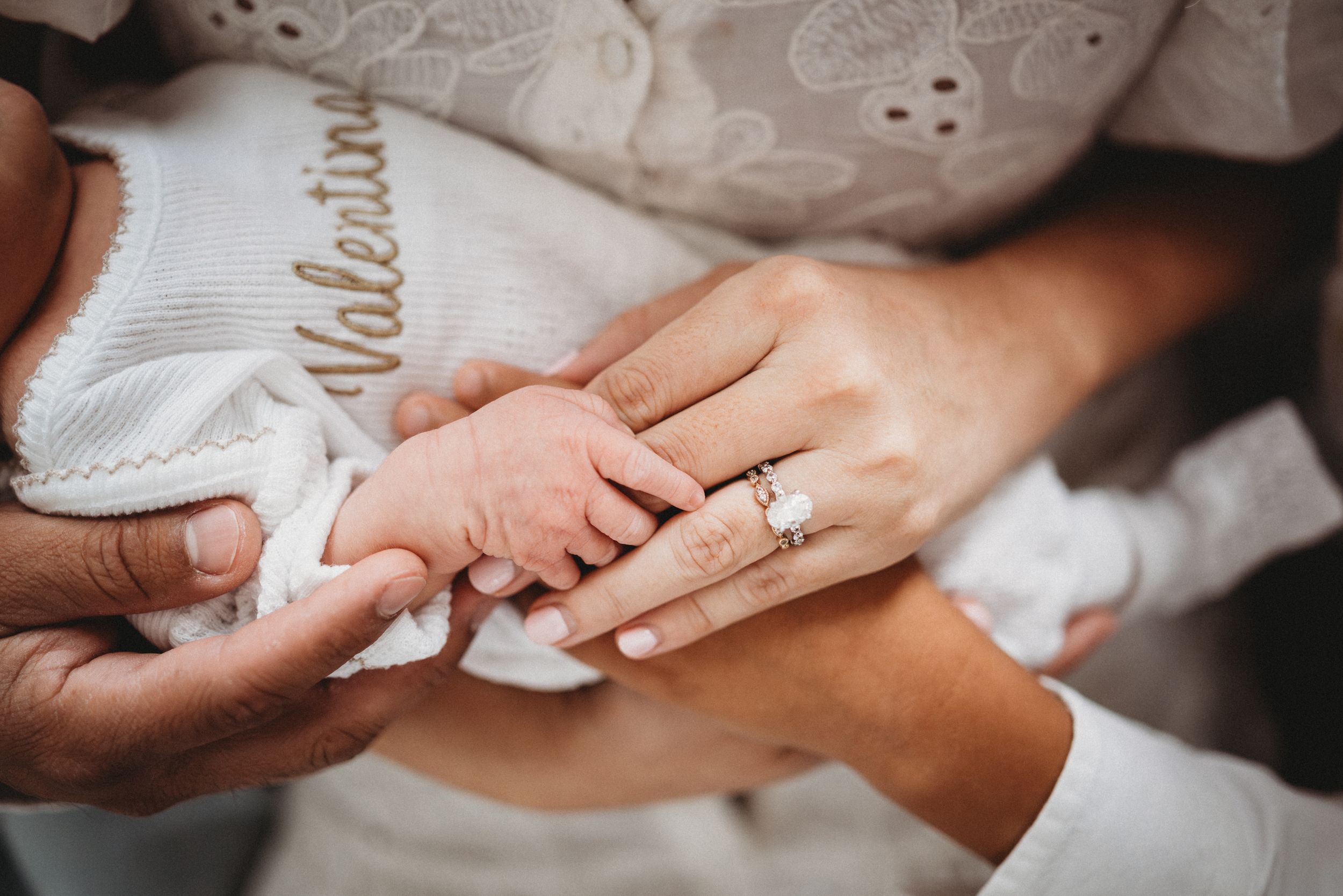 Newborn baby named Valentina holding parents hands, mother wearing diamond engagement ring, tender family moment.