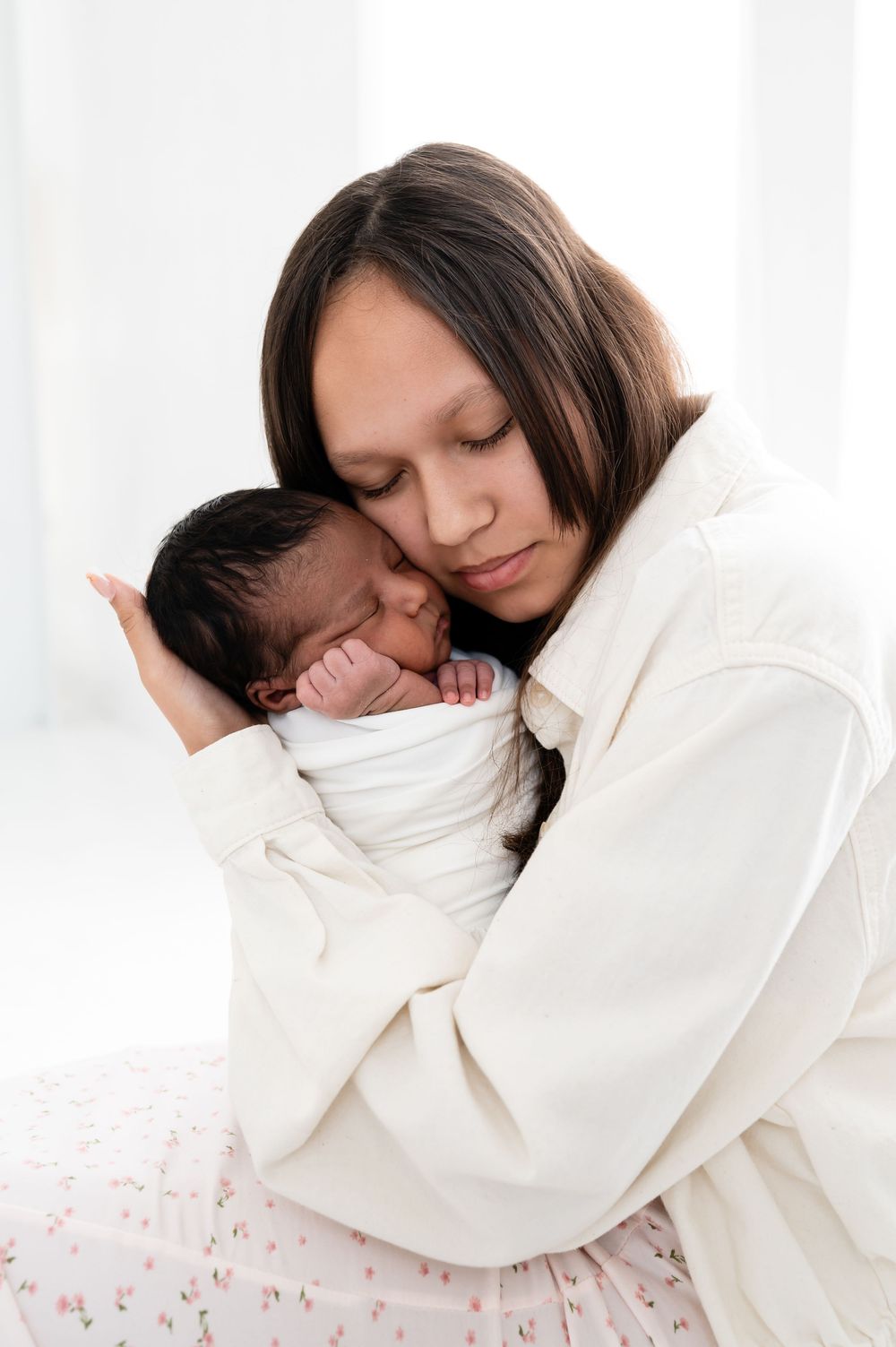 A mother tenderly holds her swaddled newborn baby close to her chest in a bright, white room.