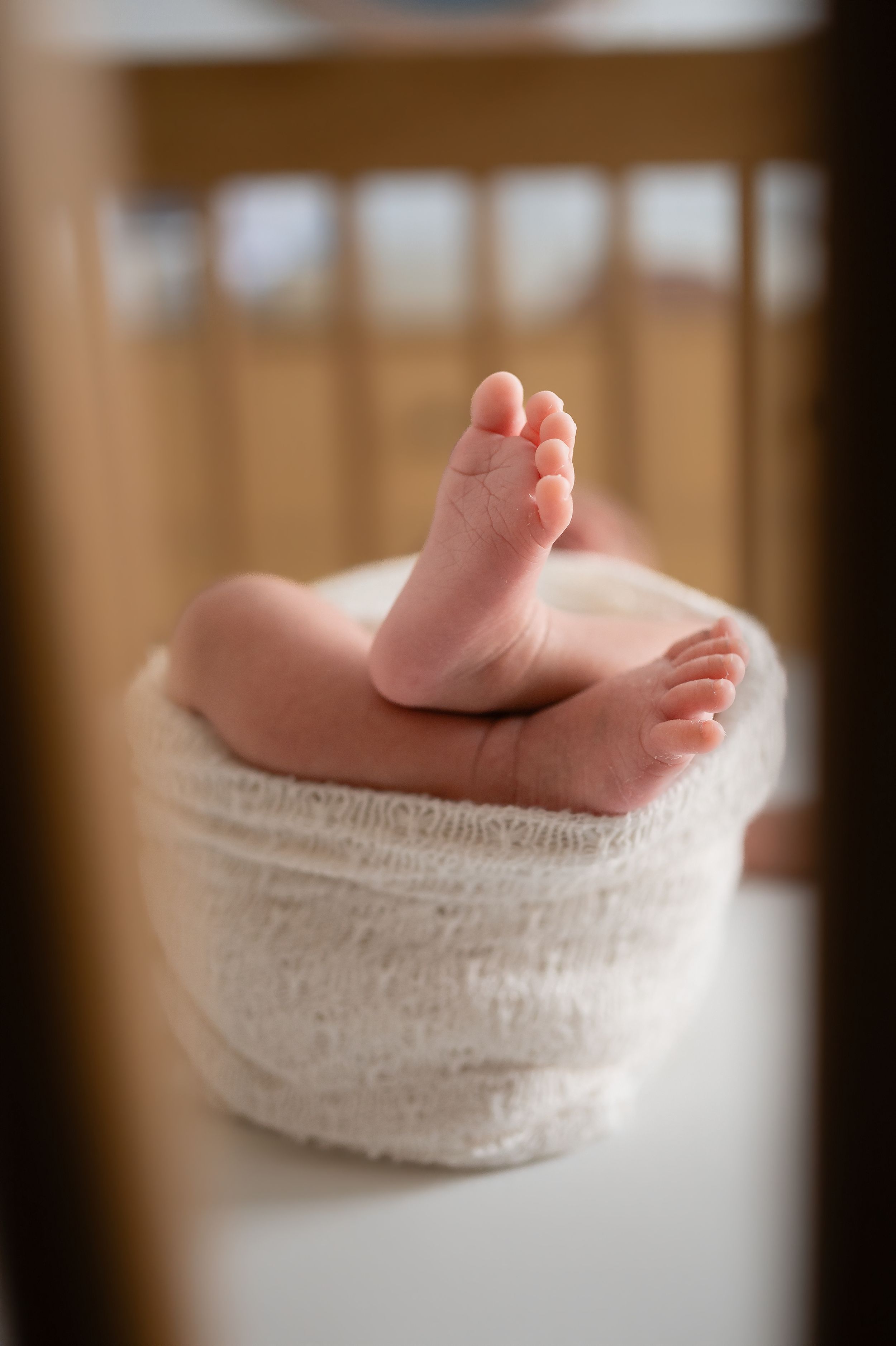 Close-up of newborn baby feet peeking out of a white swaddle blanket in a wooden crib.