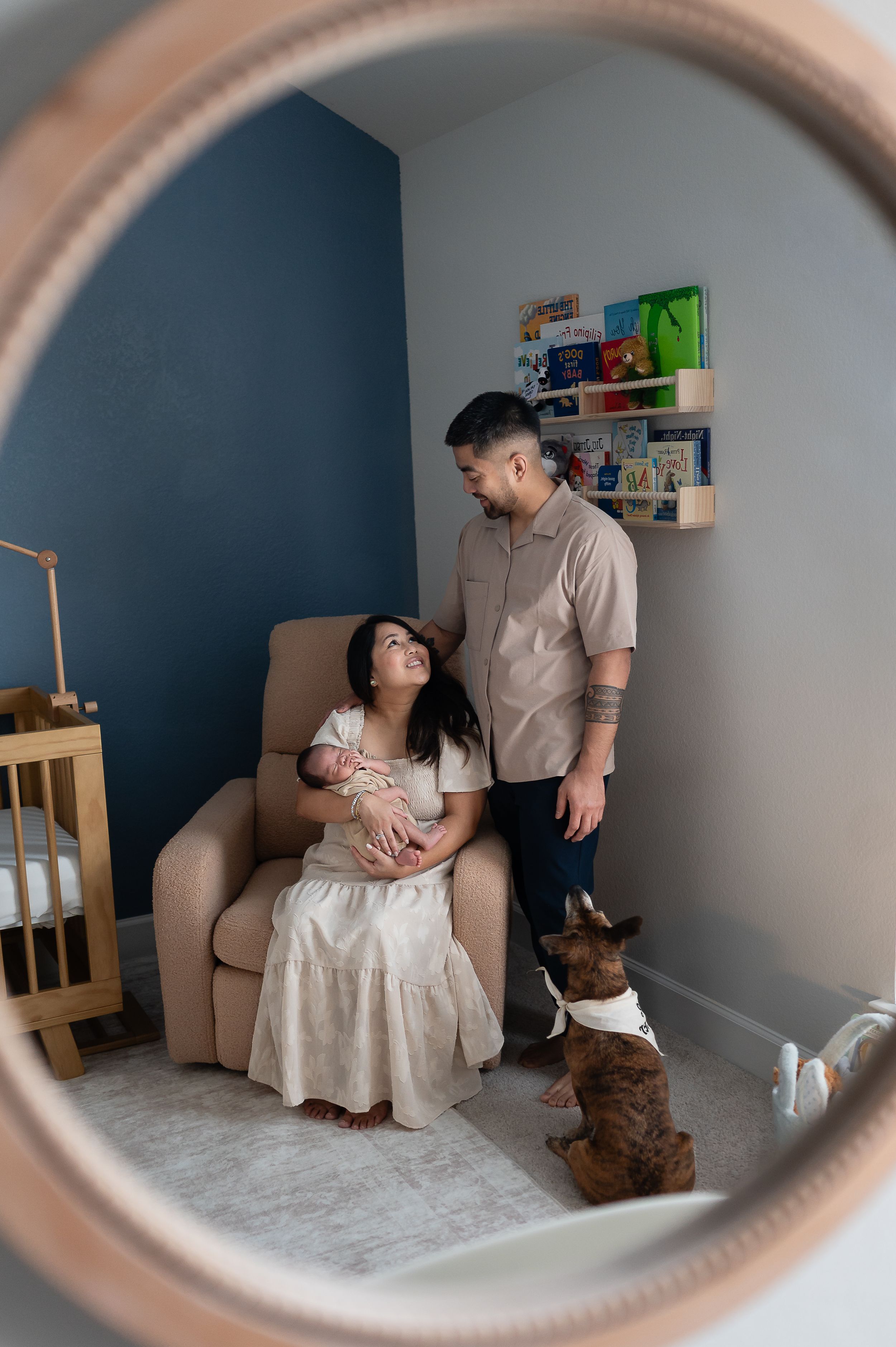 New parents sit and stand in a nursery, holding their newborn baby, with their dog nearby, reflected in a round mirror.