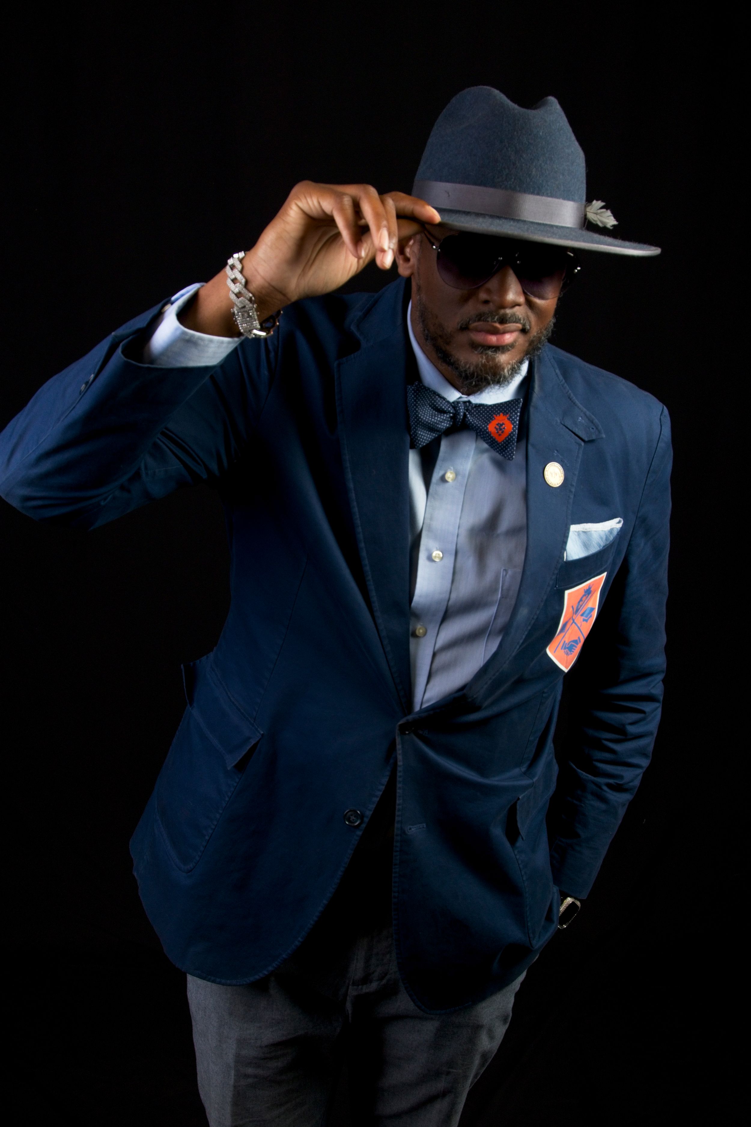 Stylish person in navy suit and fedora poses against black background.