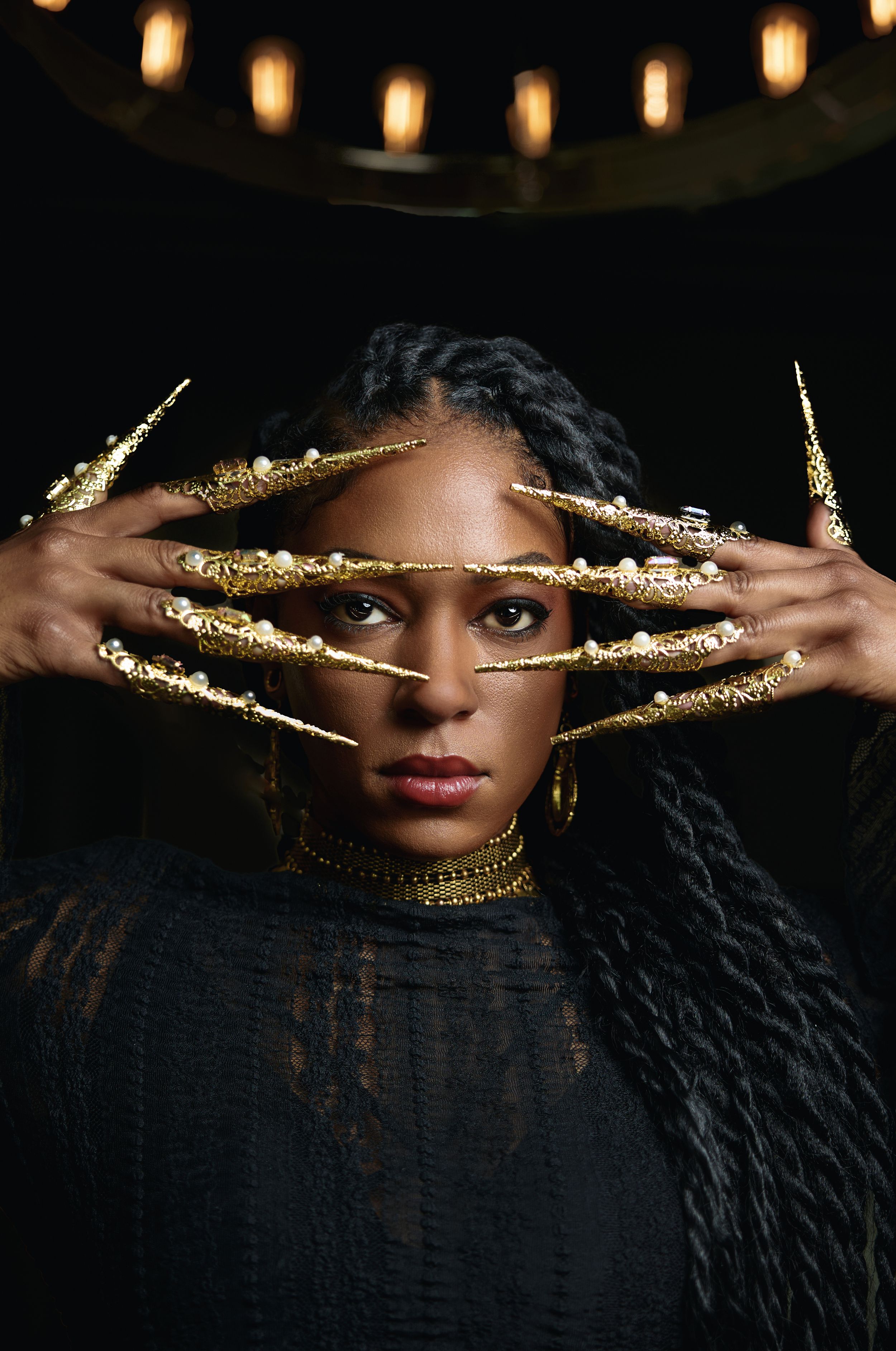 Creative portrait with gold-painted hands framing face against dark background with hanging lights.