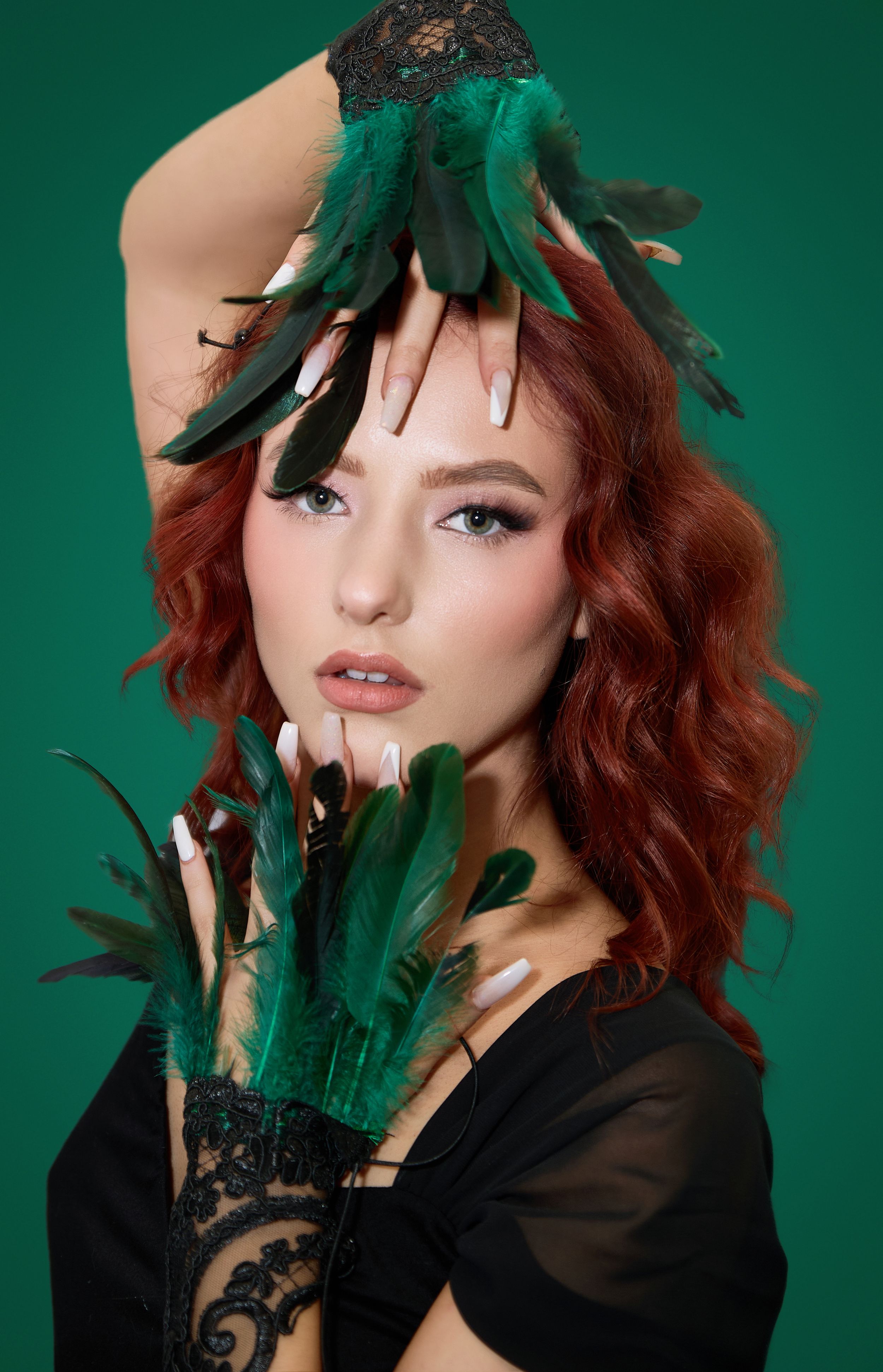 Fashion portrait with emerald green feather headpiece and black lace gloves against dark background.