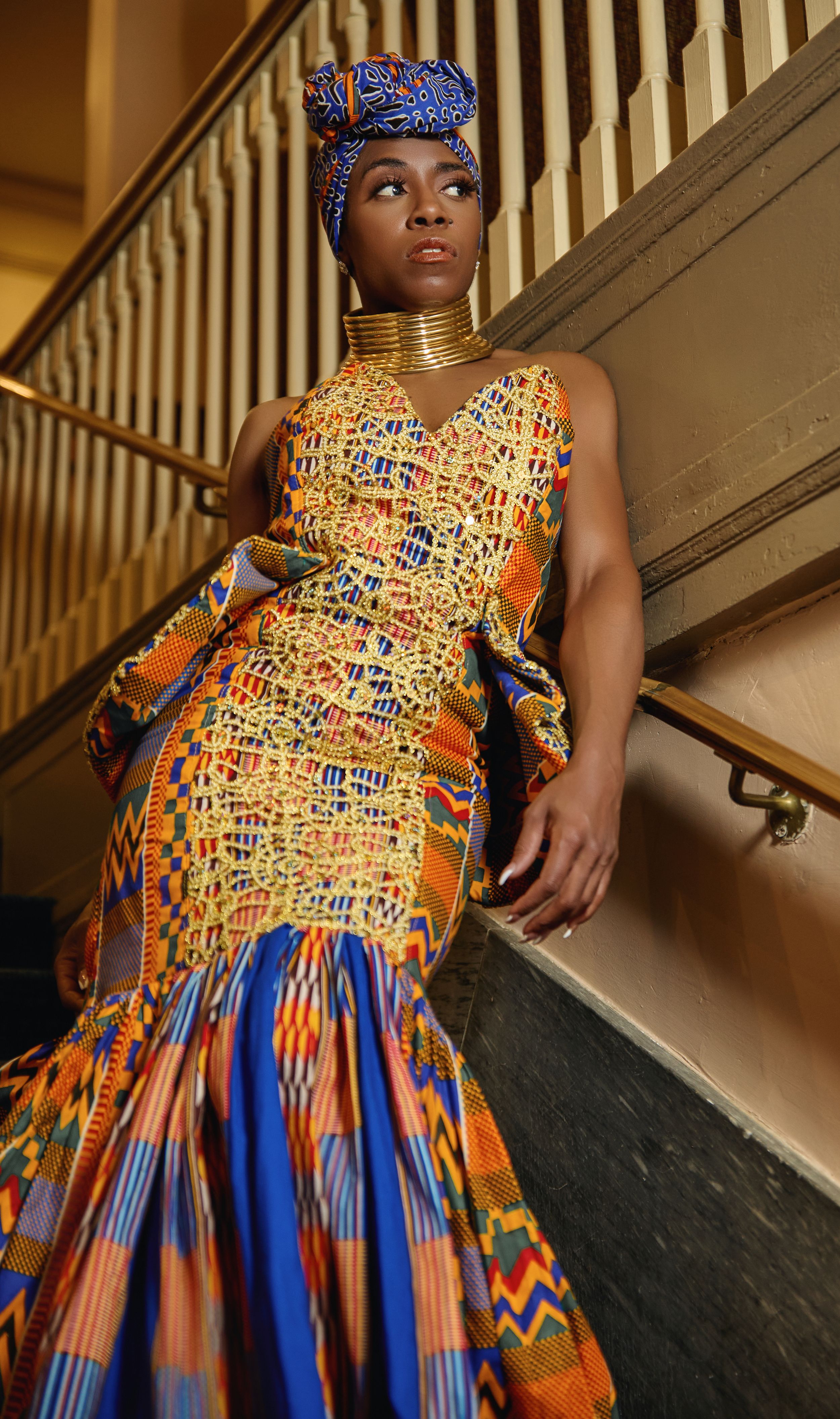 Model in vibrant African print mermaid gown and headwrap poses on elegant staircase.