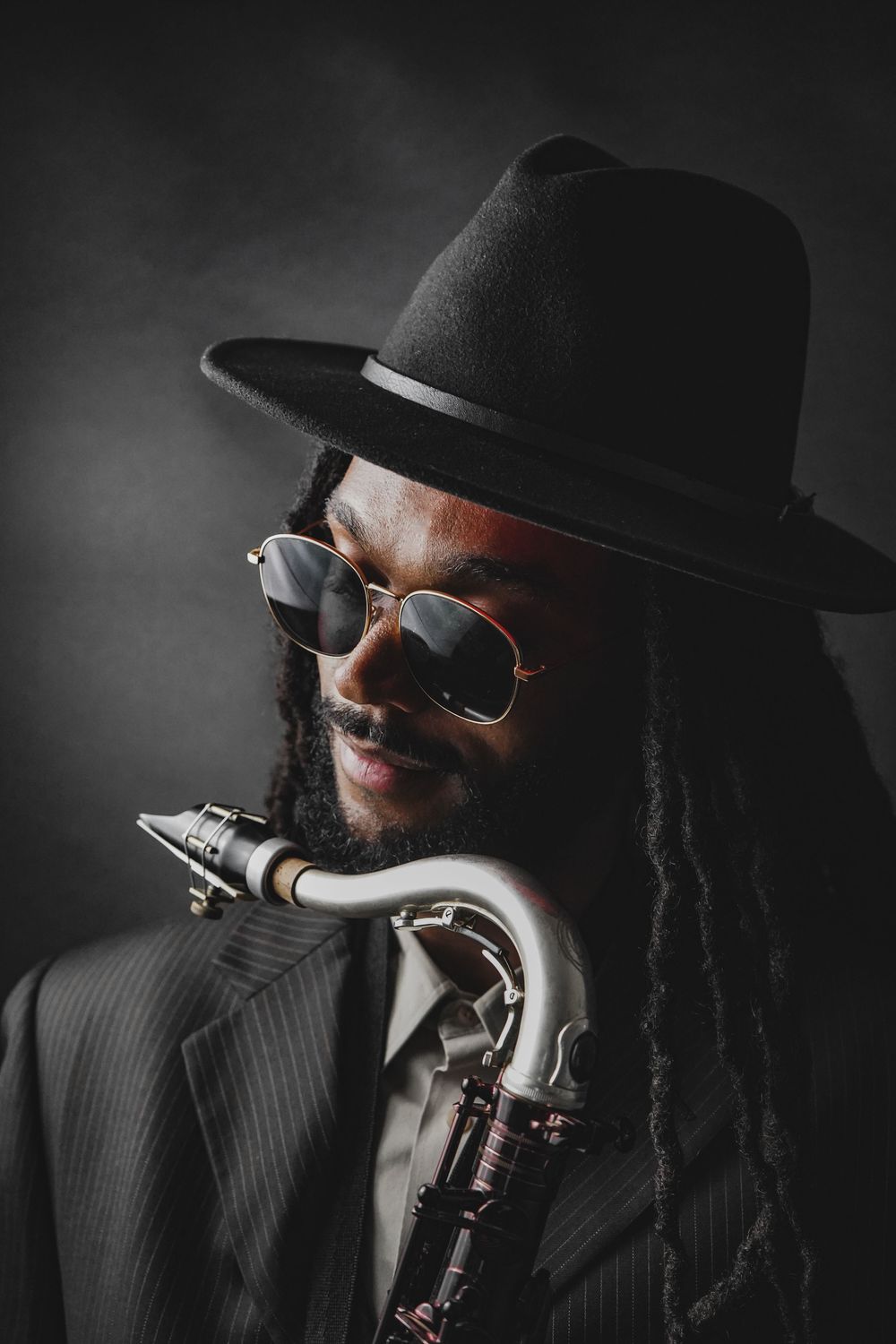Jazz musician with dreadlocks wearing a black fedora, sunglasses, and suit, holding a saxophone in a moody studio portrait.