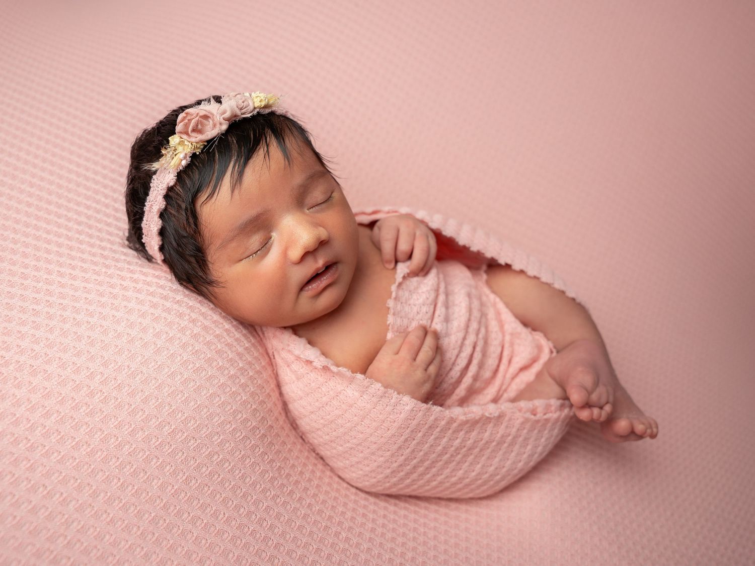 Baby girl with long dark hair, wrapped in a pink textured fabric on a pink backdrop backdrop at newborn photoshoot in Doncaster.