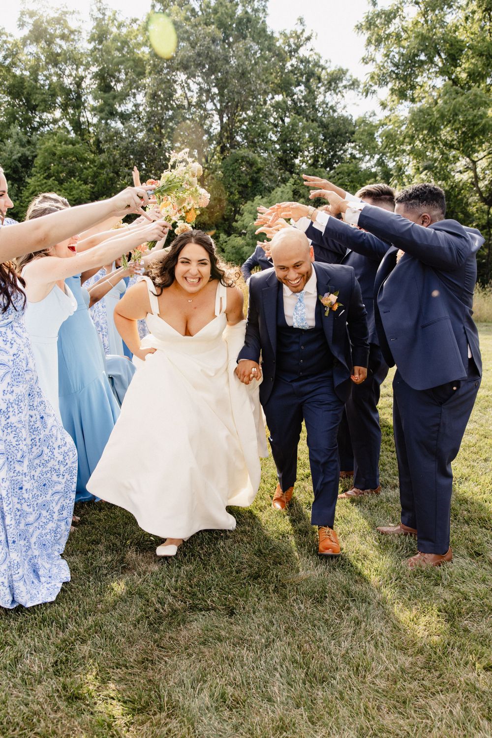 Joyful wedding party poses in a line on grass with bridesmaids in blue dresses and groomsmen in navy suits.