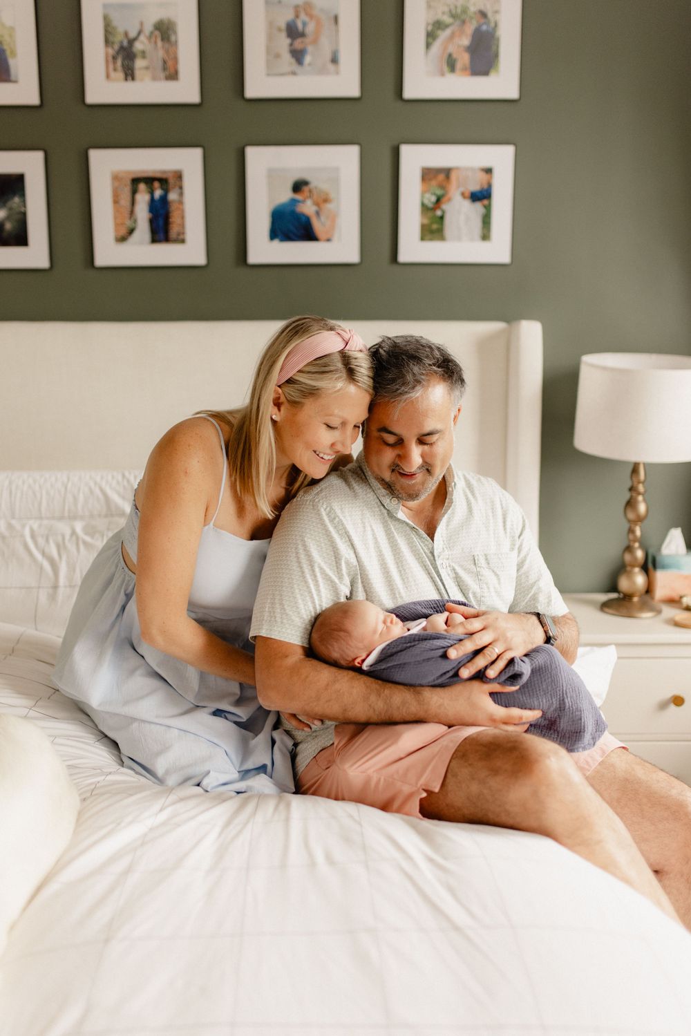 Couple on white couch holding newborn baby in cozy bedroom with framed photos on olive green wall.