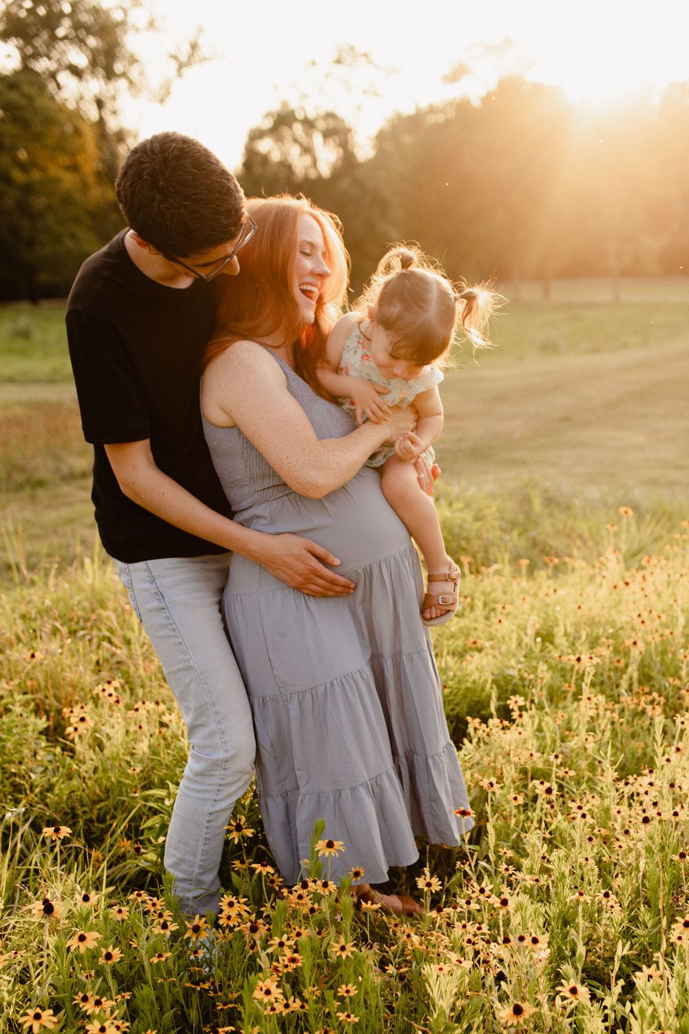 Family embracing in golden sunset field with expecting mother in flowy dress.