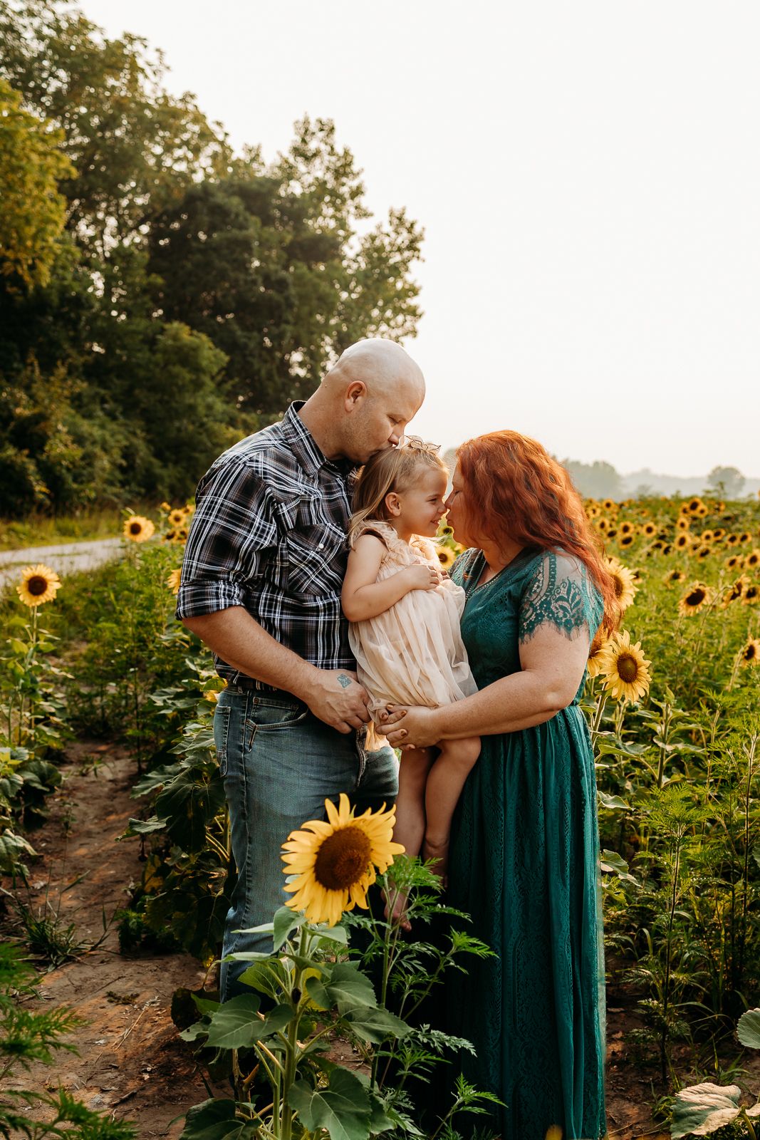 A mom, dad, and little girl all hugging each other. The dad is kissing the little girl on the head. They are all standing in sunflowers.