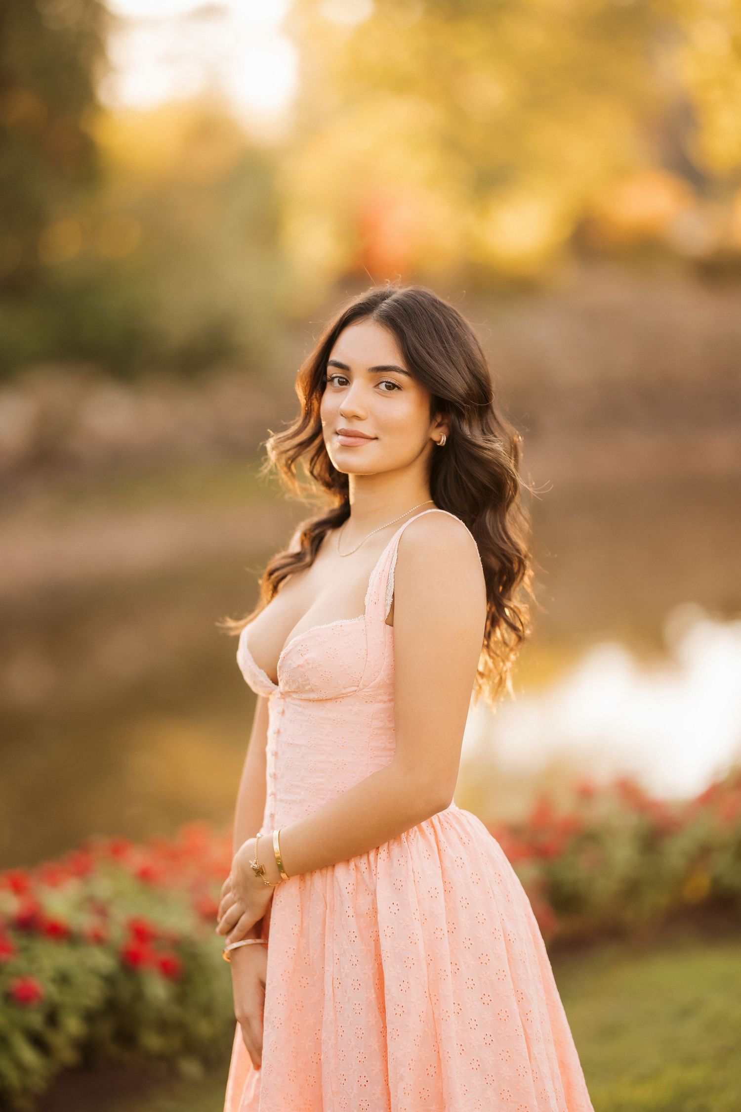 A person in a peach-colored dress poses outdoors during golden hour with a lake and autumn foliage in the background.