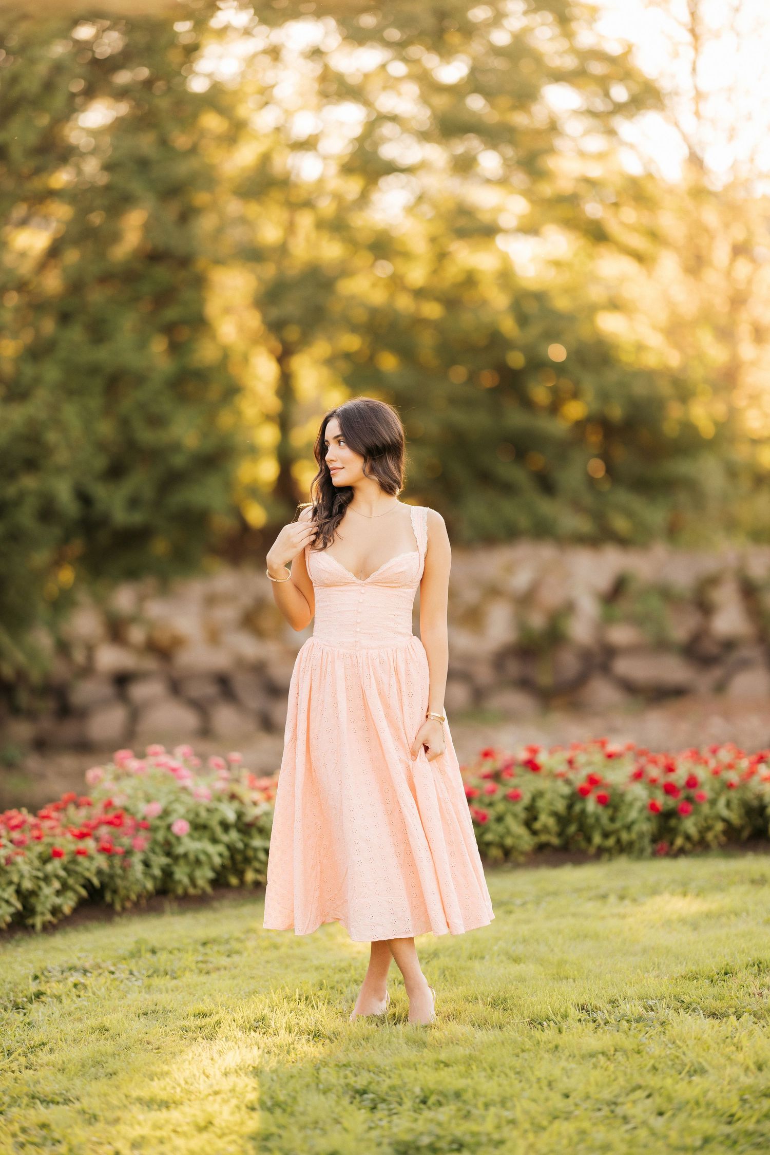 A person in a peach-colored dress stands barefoot on grass near a garden with red flowers at sunset.