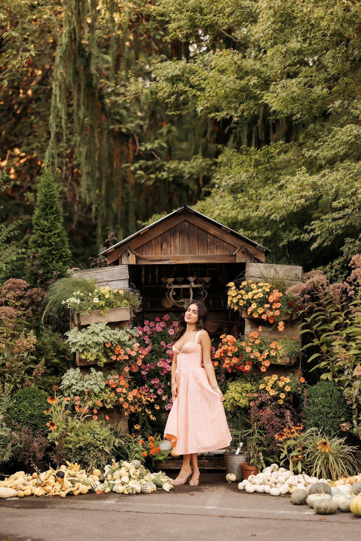 A dreamy garden scene with a pink dress against a rustic wooden shed surrounded by colorful flowers and white pumpkins.