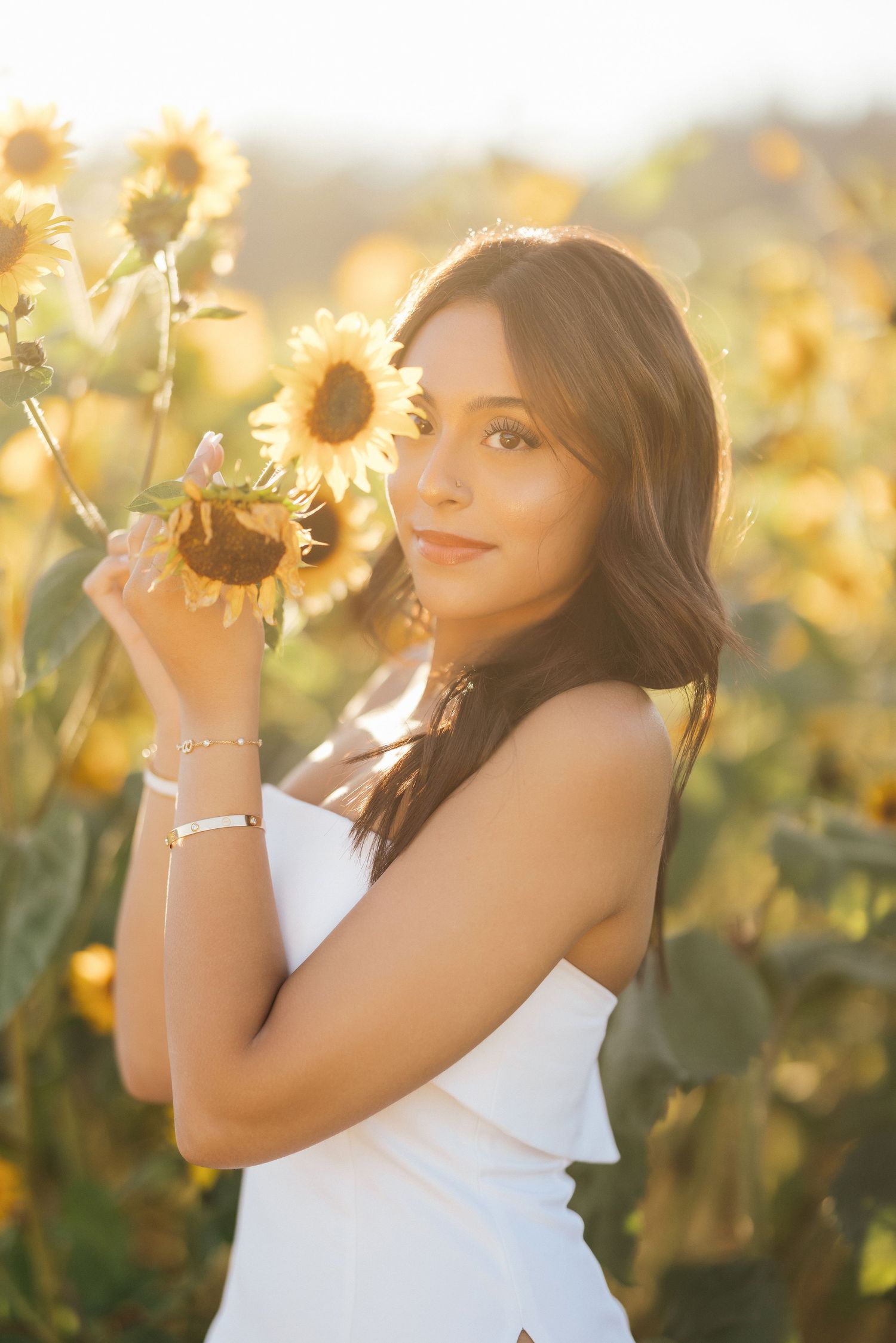 A woman in a white dress poses with sunflowers in a golden-lit field during sunset.