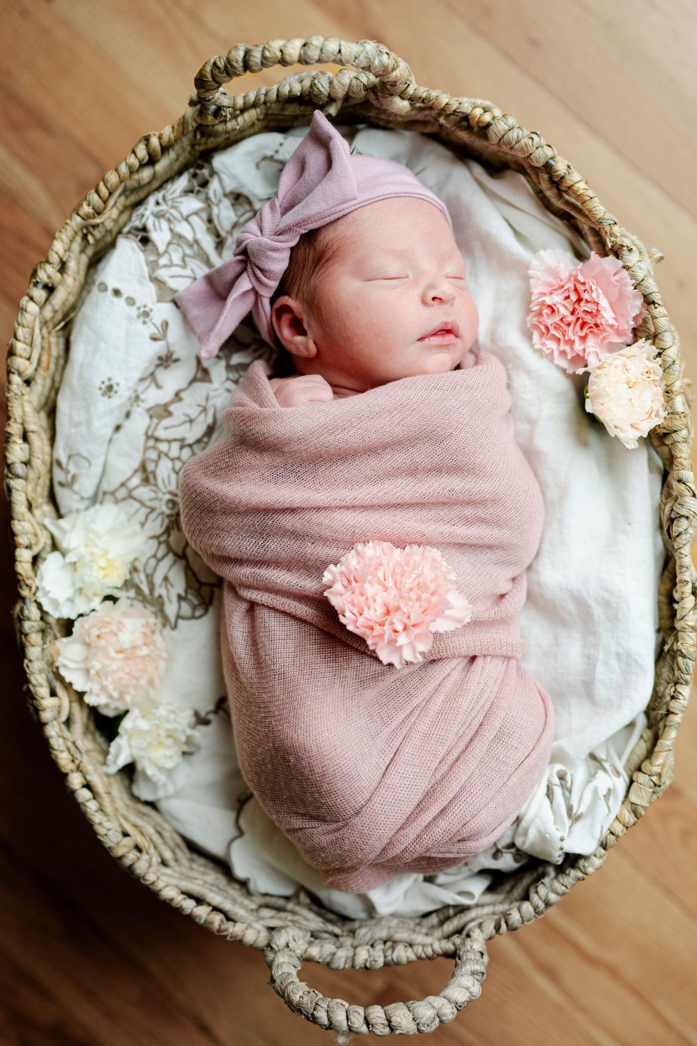 Sleeping newborn baby swaddled in pink wrap with floral headband resting in a woven basket with flowers.