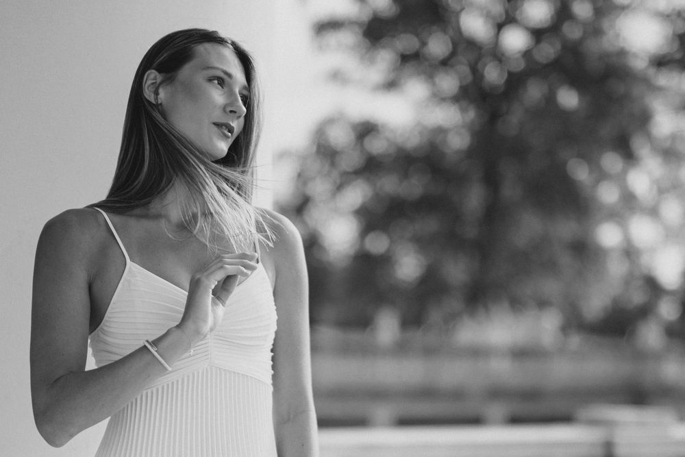 Black and white portrait of a young woman in a white dress, gazing sideways with blurred trees in background.