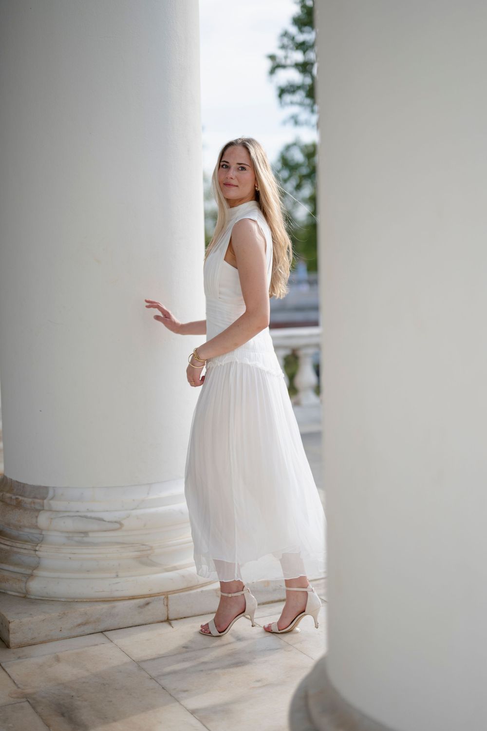 Young blonde woman in elegant white pleated midi dress and heels posing between white columns.
