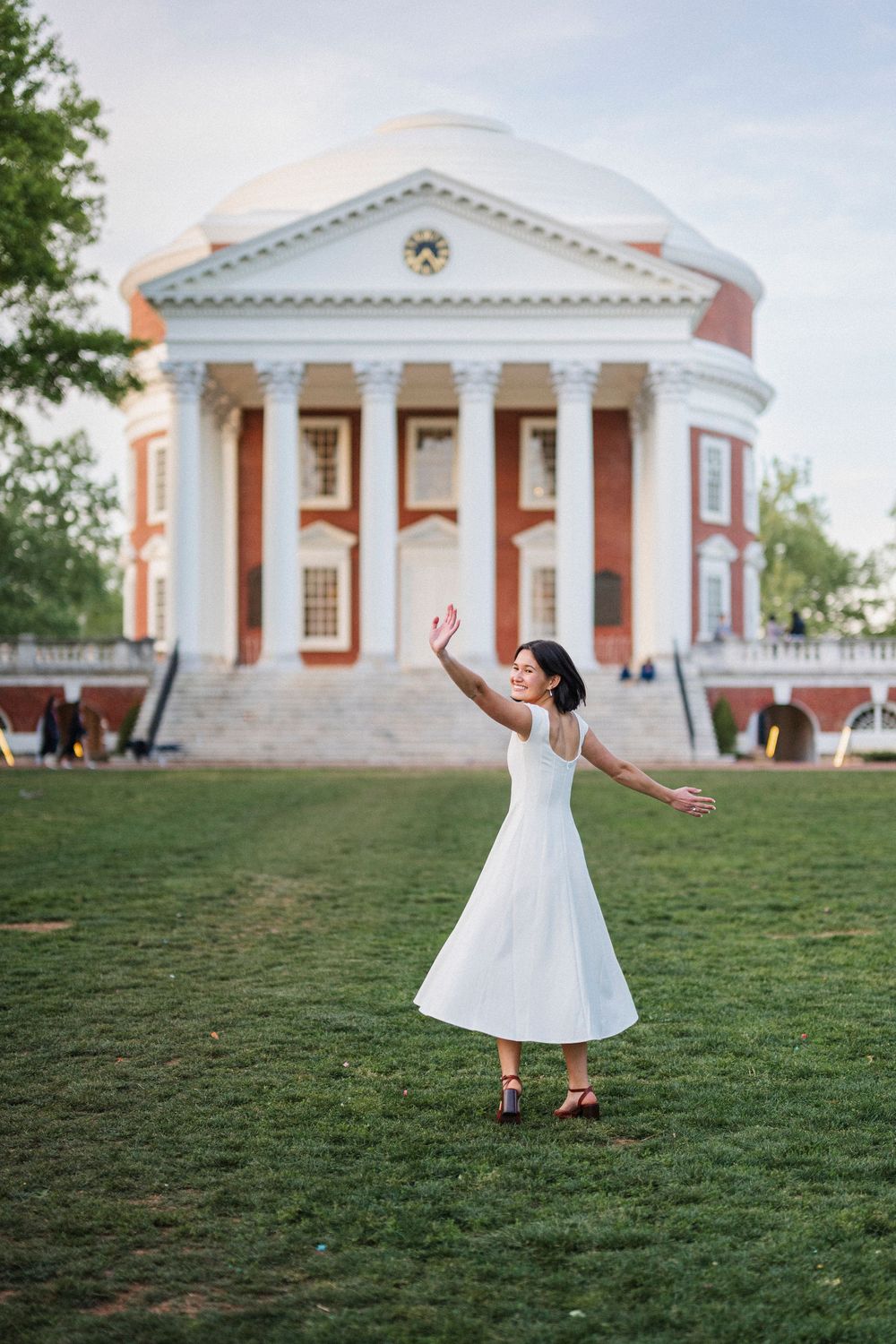 Woman in white dress dancing on green lawn before a grand neoclassical building with red brick and white columns.