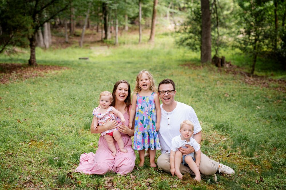 Happy family of five smiling together on green grass in a wooded park setting during a outdoor photo session.