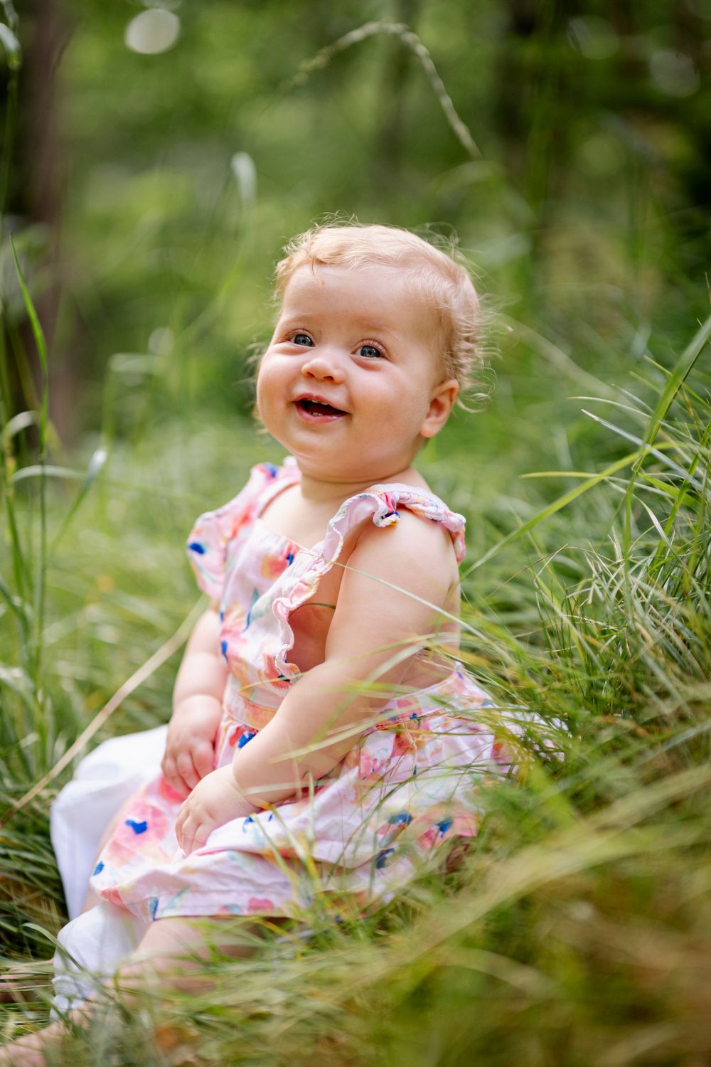 A happy baby girl in a pink floral dress sitting in tall green grass outdoors, smiling joyfully.